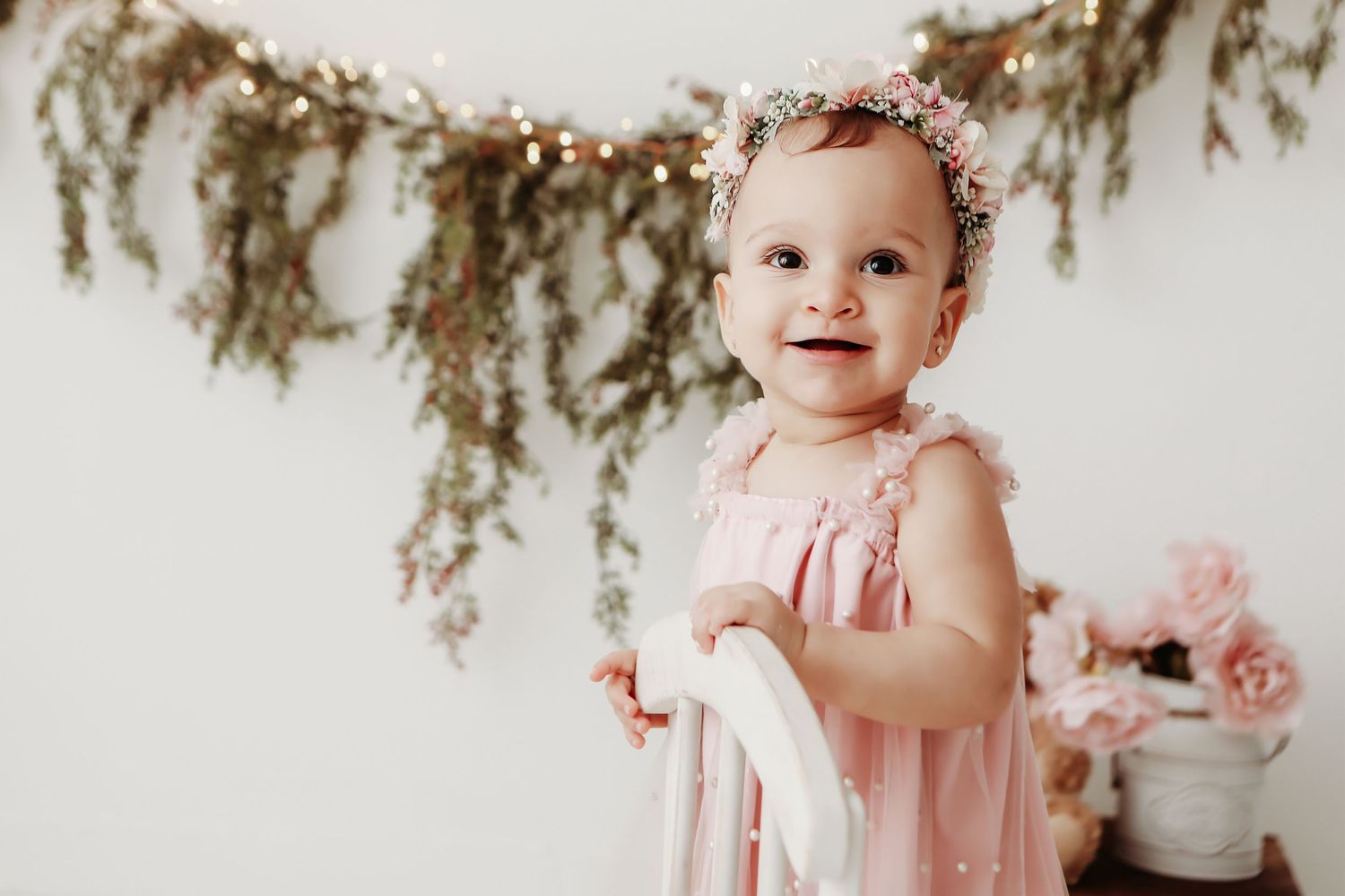 A baby in a pink dress sits on a white backdrop surrounded by floral decorations and rustic garlands.