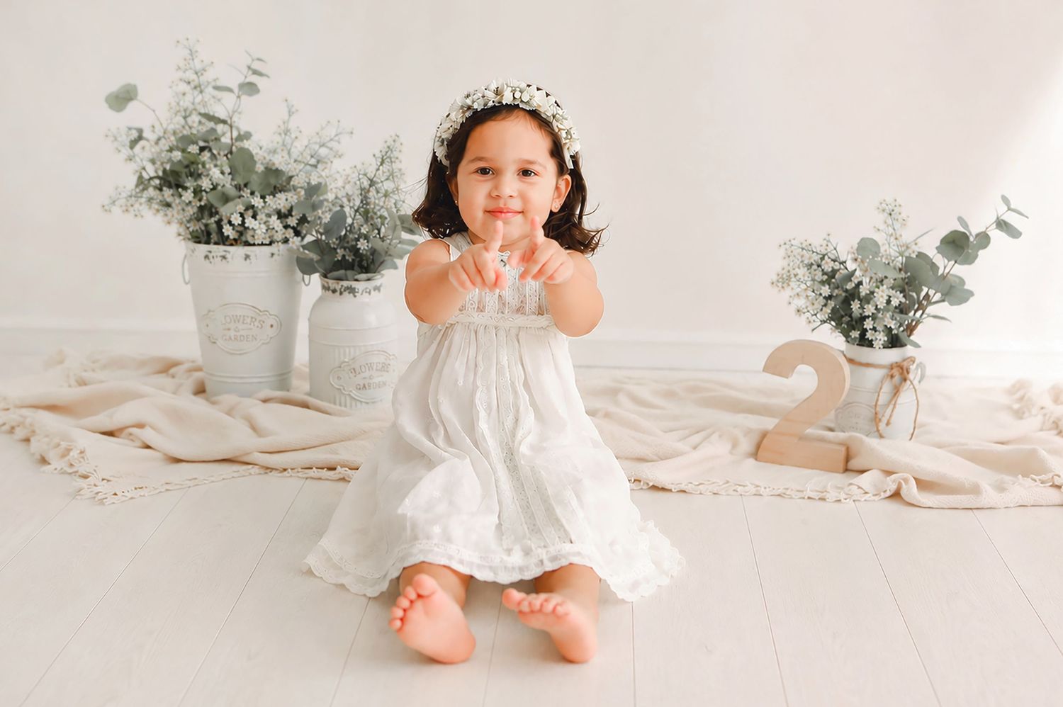 Toddler girl in white lace dress and floral crown sits on floor with greenery decor for second birthday photo.
