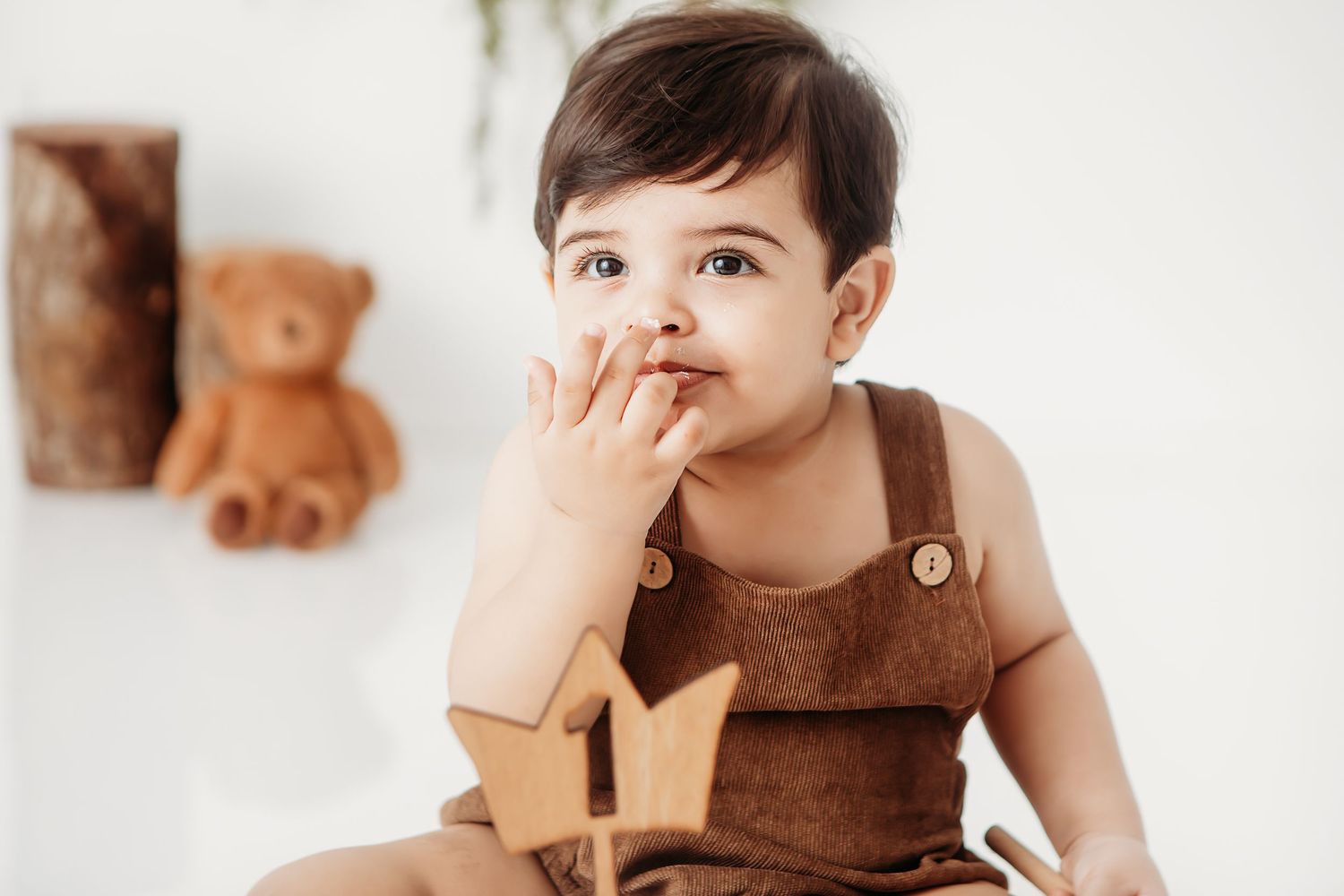 Small child in brown overalls sits against white wall with teddy bear decor in background.
