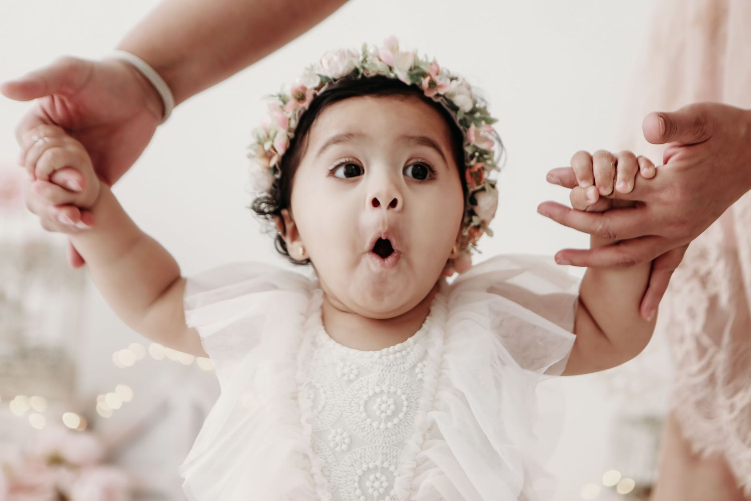 Baby wearing white lace outfit and flower crown reaches up playfully during photo session.