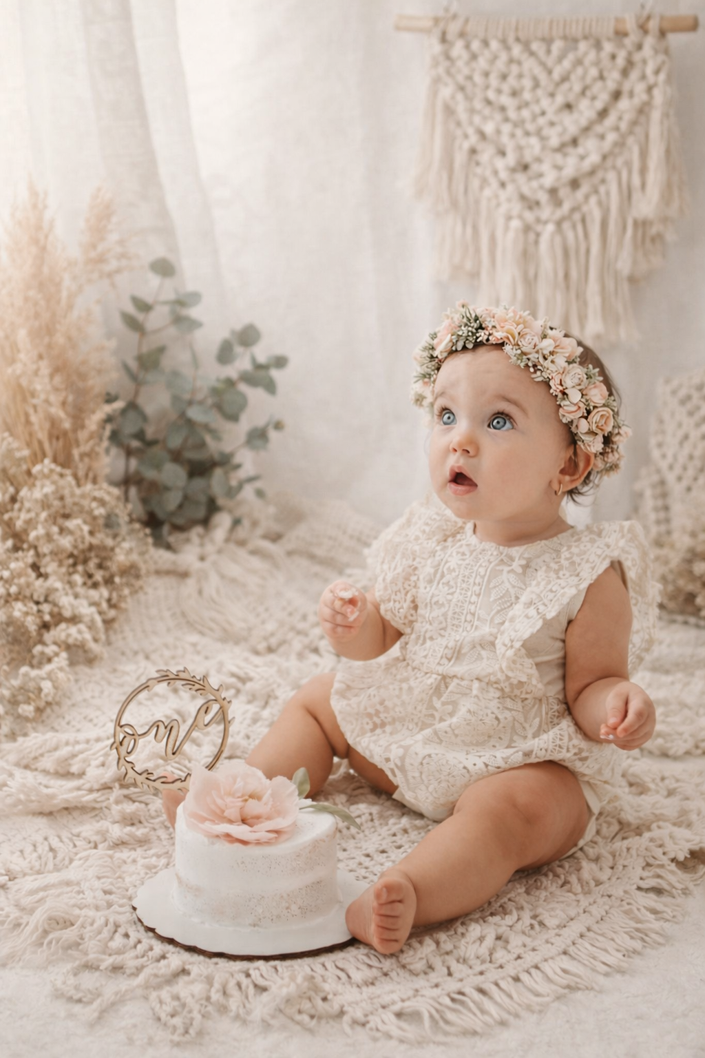 A baby in a lace dress and floral crown sits on cream macrame with vintage decor and pampas grass in a dreamy setting.
