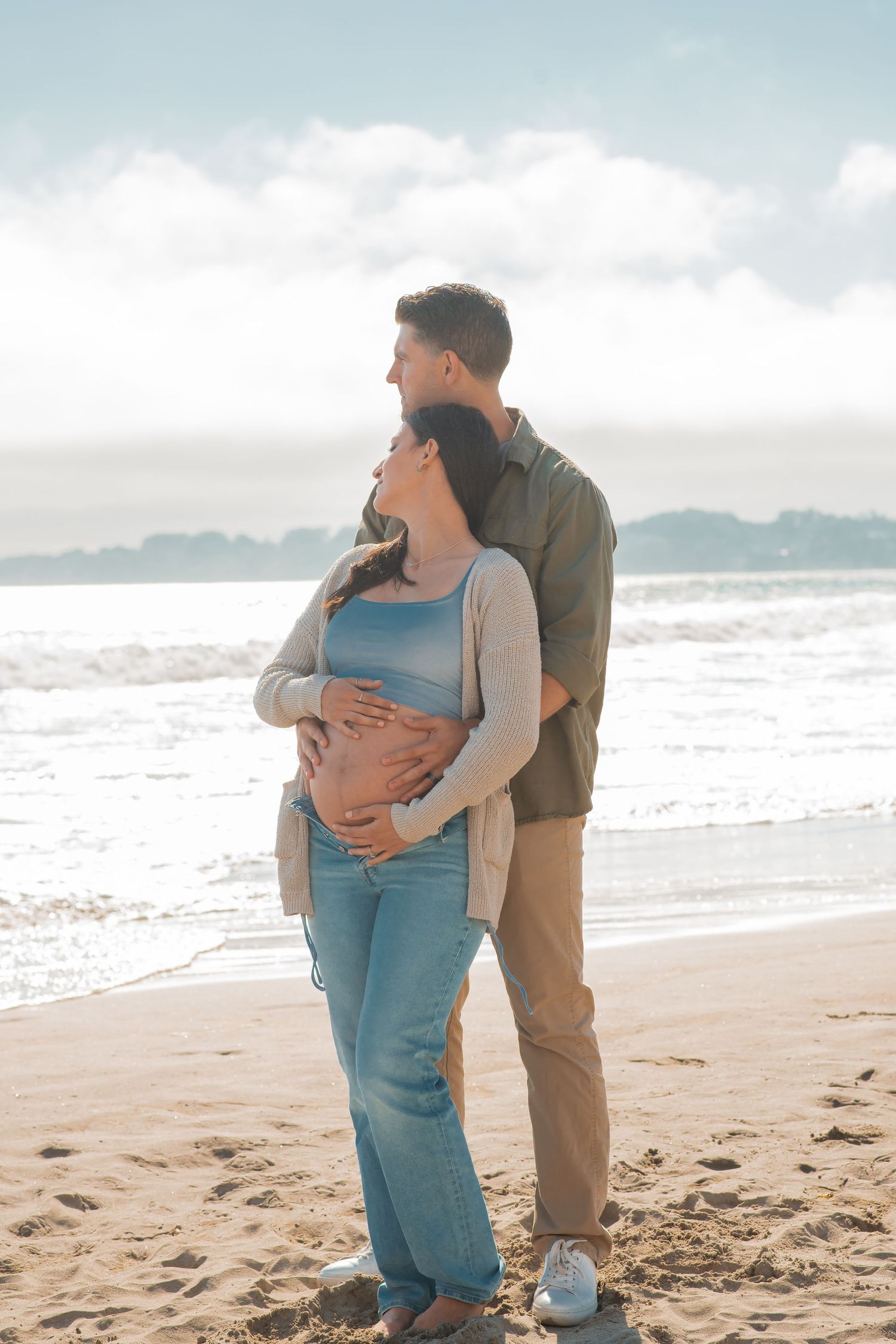 A couple embraces tenderly on a beach during a maternity photoshoot, with gentle waves in the background.