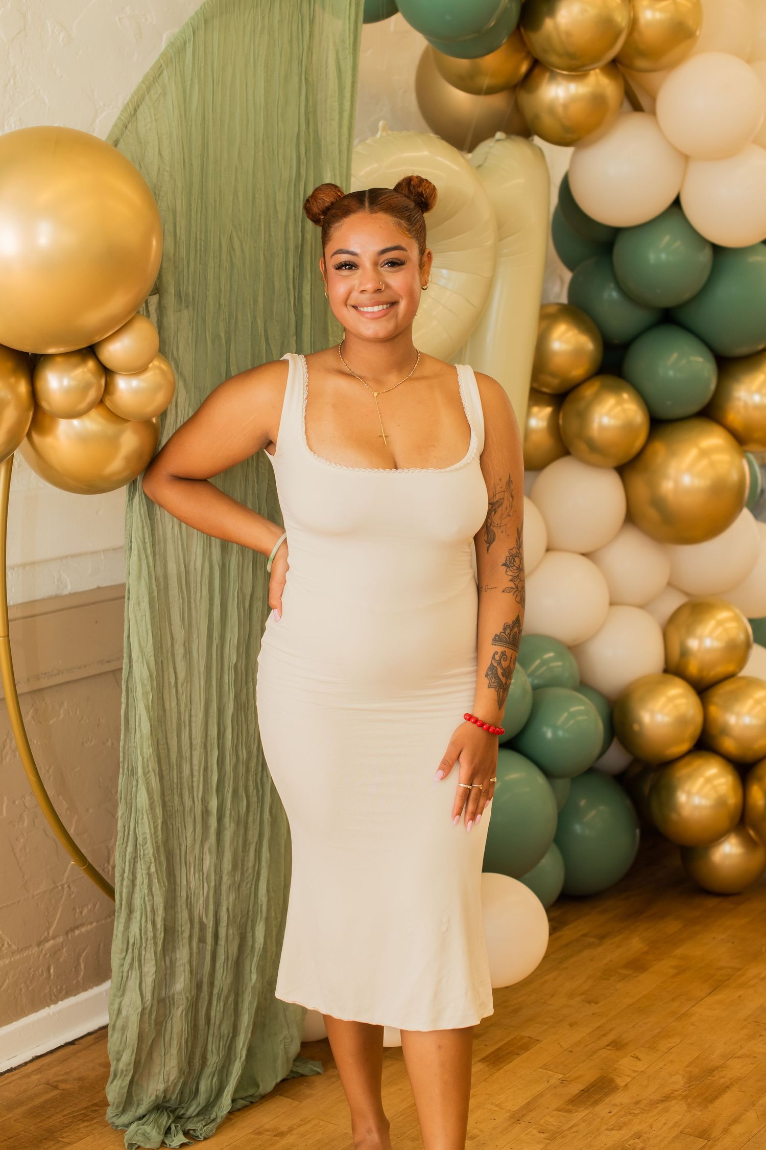 A festive party setup with sage green draping and gold and white balloon arch against a wall with hardwood floors.