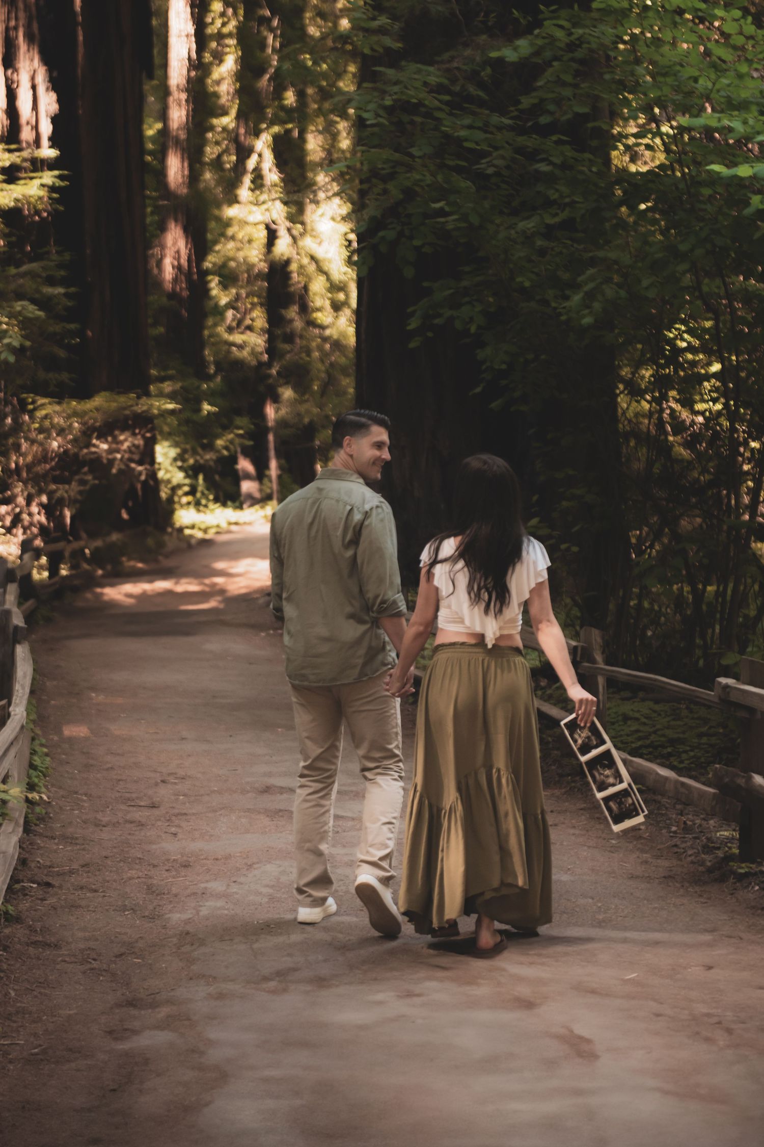 A couple walks together on a serene forest path while one carries a wooden box on a sunny afternoon.