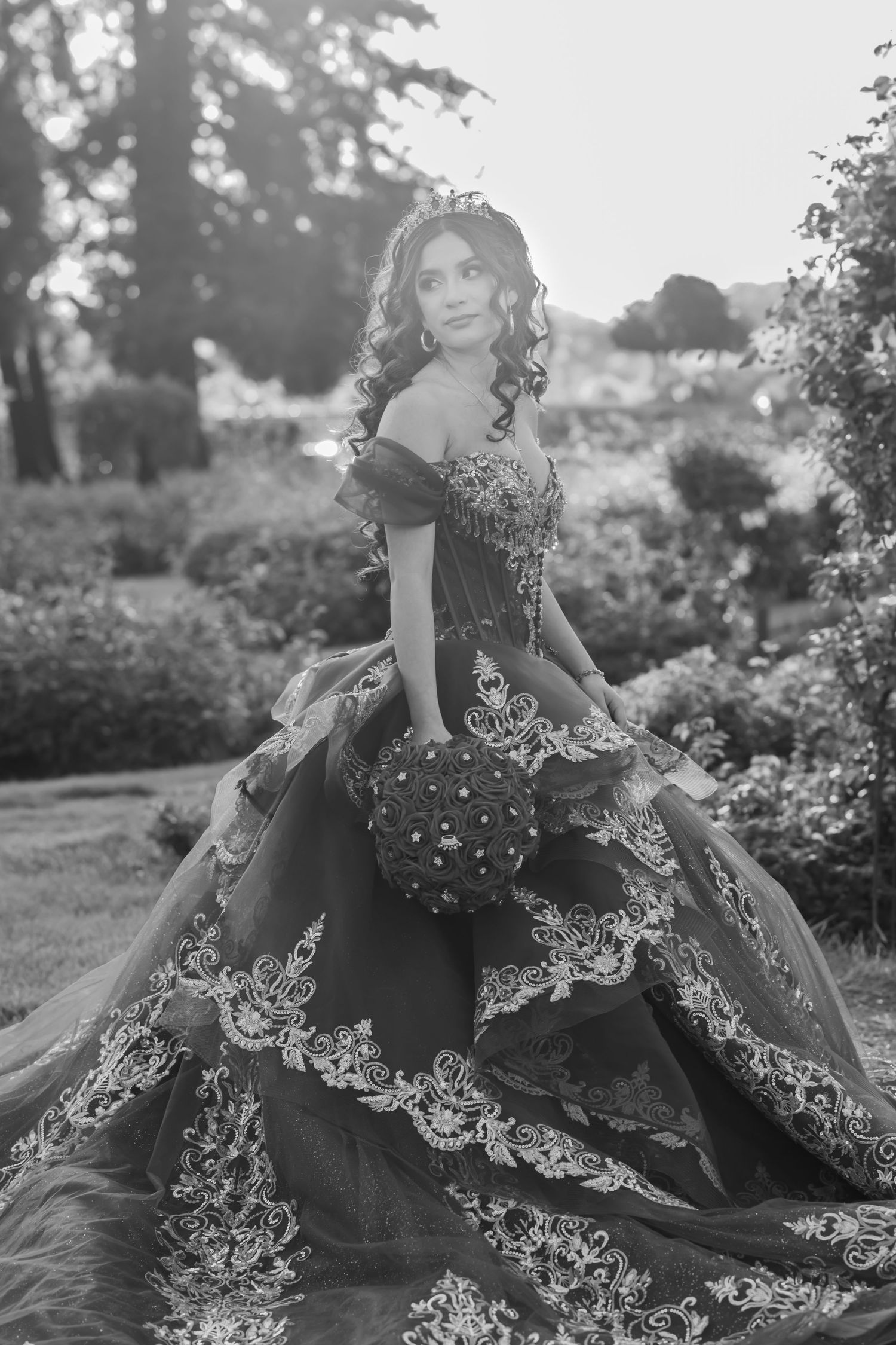 Black and white photo of an elegant ball gown with intricate lace details and off-shoulder design in a garden setting.