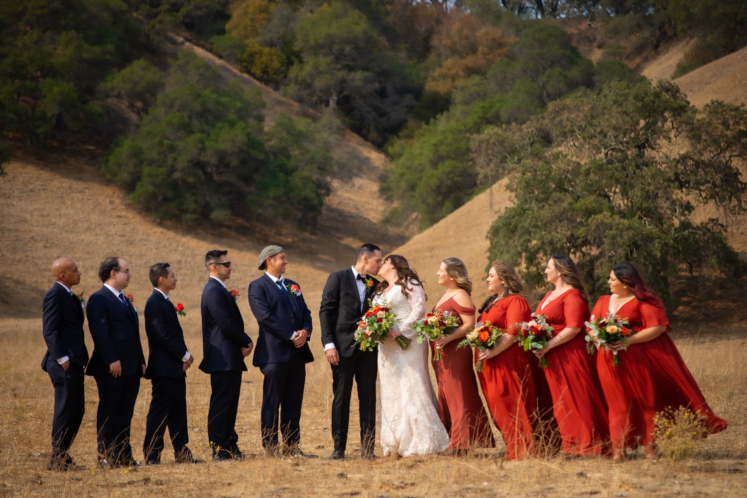Wedding party in dark suits and vibrant red dresses pose together on a hillside with autumn trees in the background.