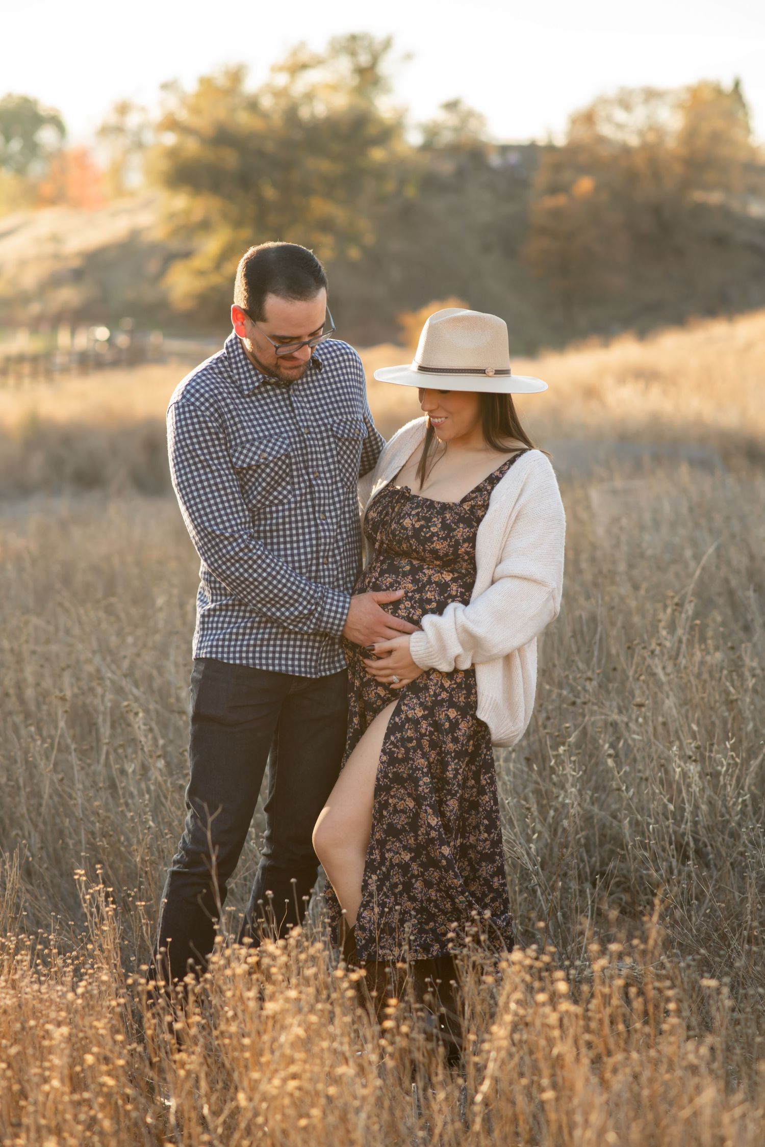 A romantic maternity photoshoot in a golden field shows a couple embracing tenderly at sunset.