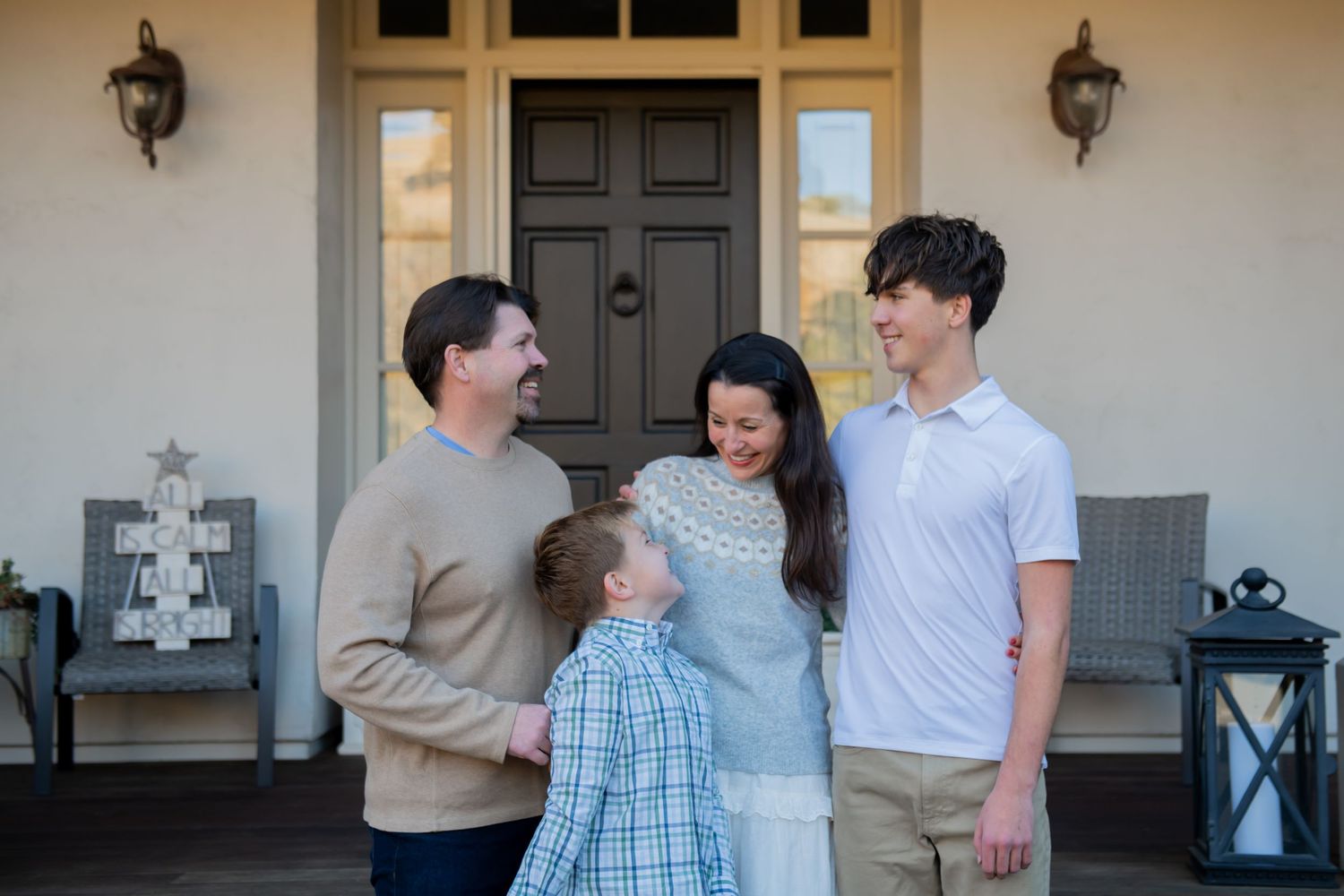 A family stands together on their porch, sharing a candid moment in front of a black door with lantern lights.