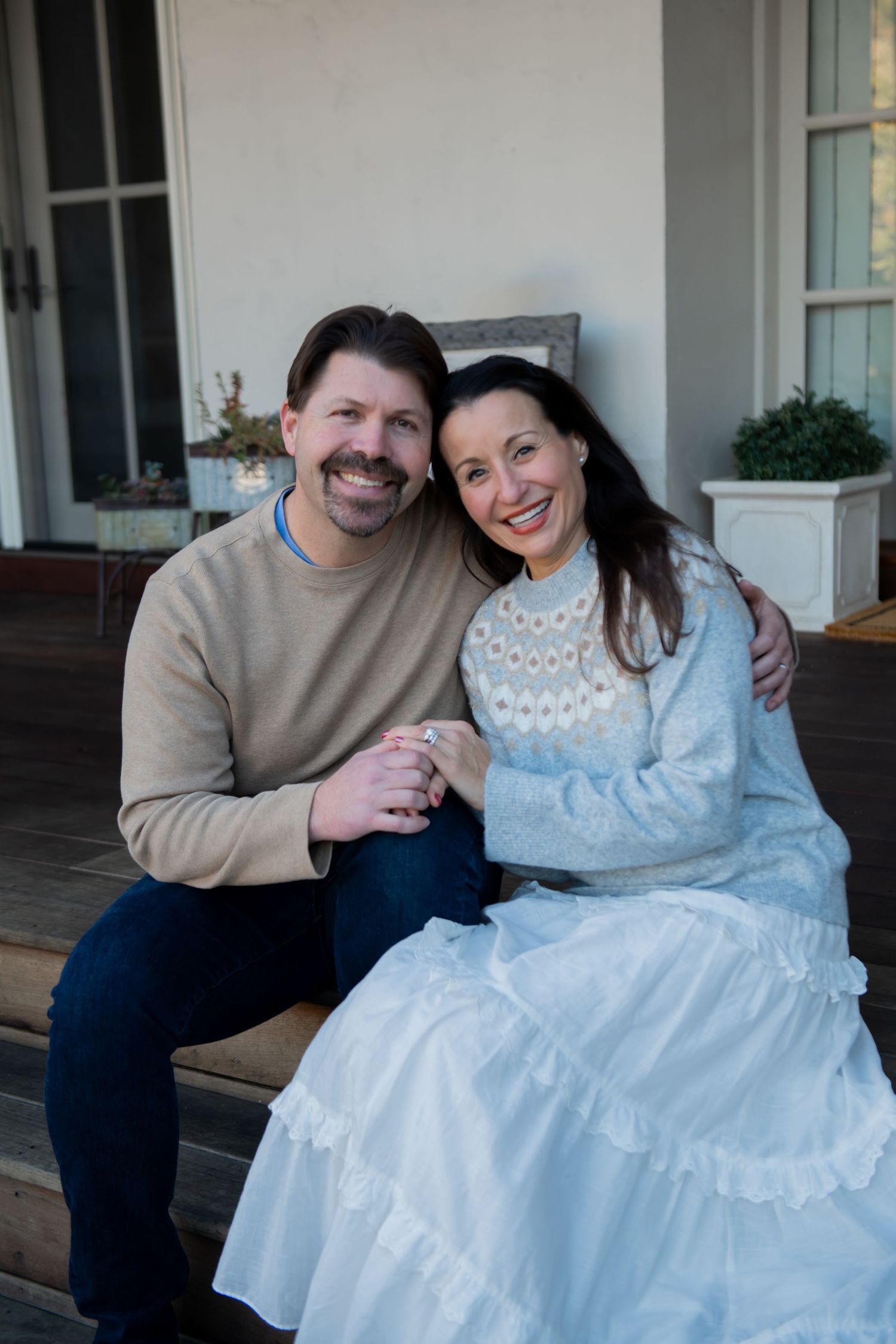 A couple sits together on a porch, with the woman wearing a blue and white dress and the man in a beige sweater.
