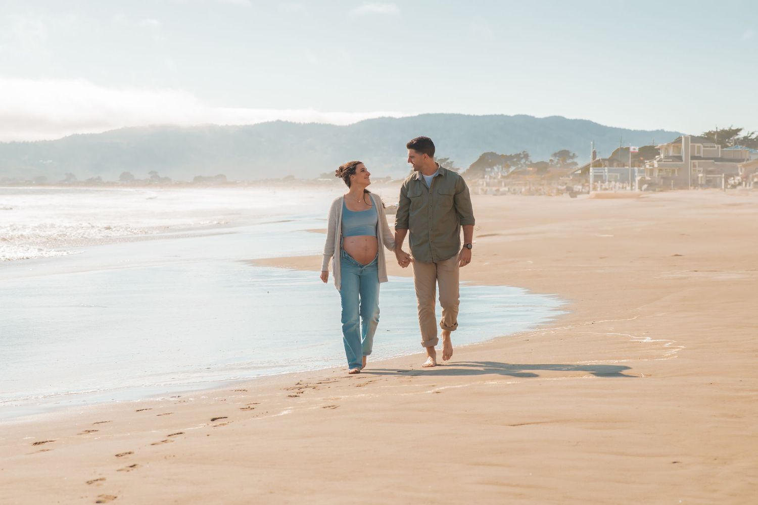 A couple walks together along a misty beach shoreline near coastal mountains on a serene morning.
