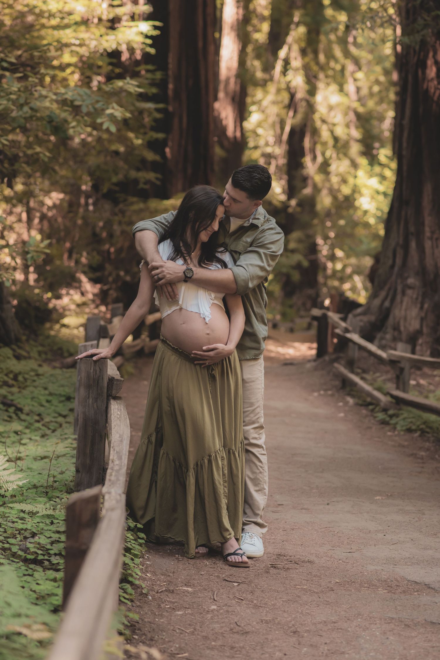 A romantic couple embraces on a sunlit forest path surrounded by tall trees and natural greenery.