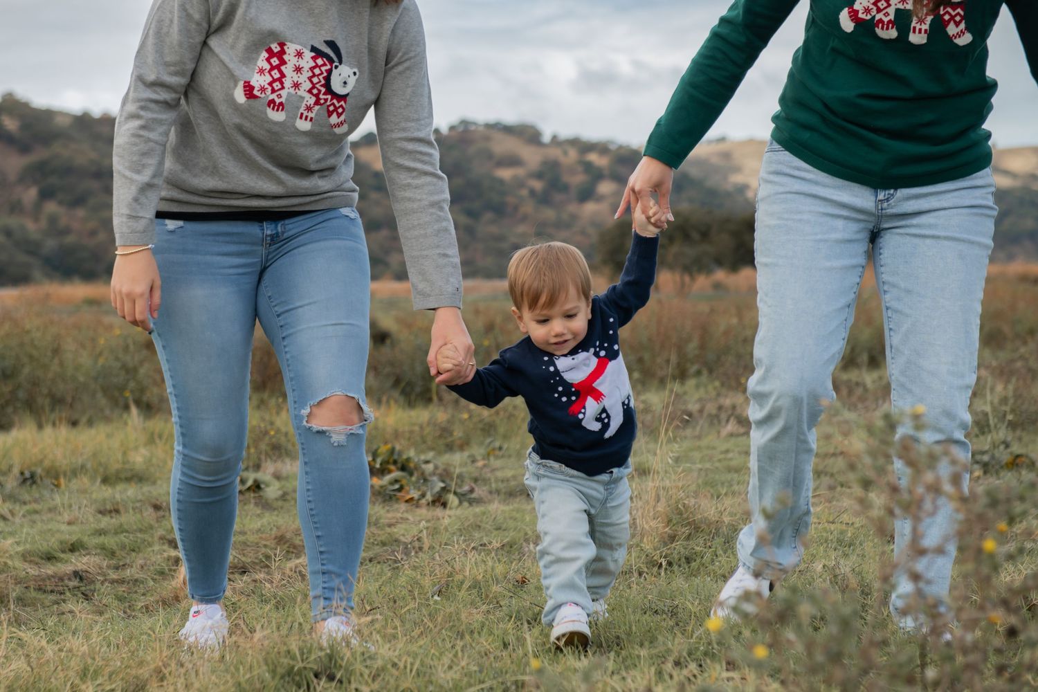 A toddler in a sweater walks holding hands with two people in jeans on a grassy hillside.