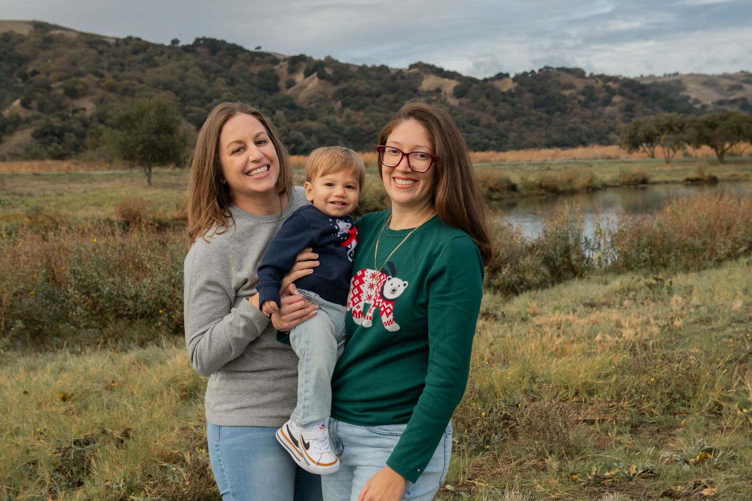 Two people and a small child pose together in front of a scenic lake and hillside on a cloudy day.