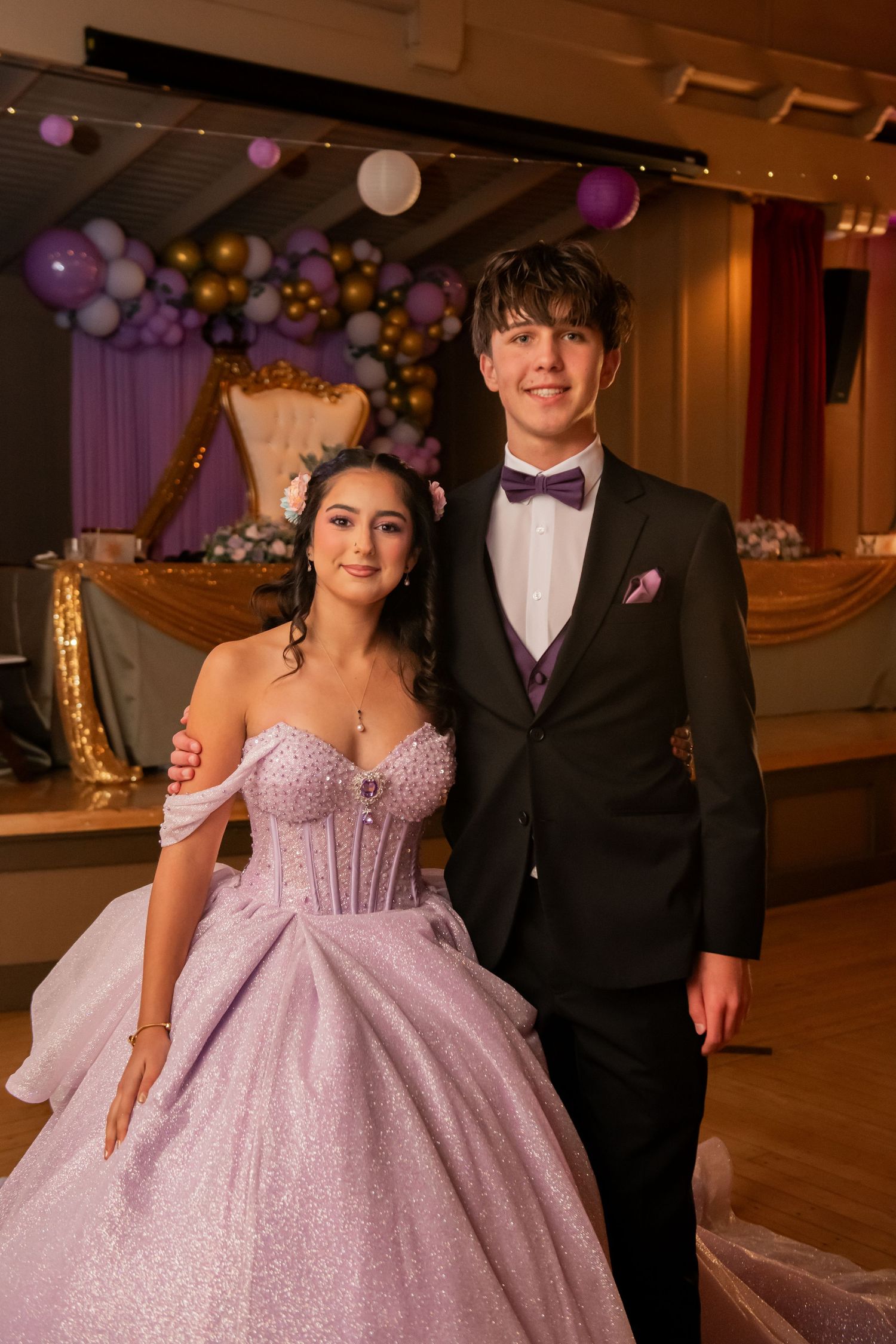 A quinceañera celebration features a grand ballroom decorated with purple and gold balloons and elegant lighting for the event.