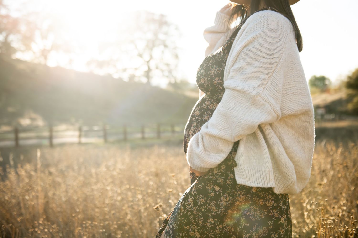 Woman in cozy cream sweater stands in sunlit golden field at sunset wearing floral dress.