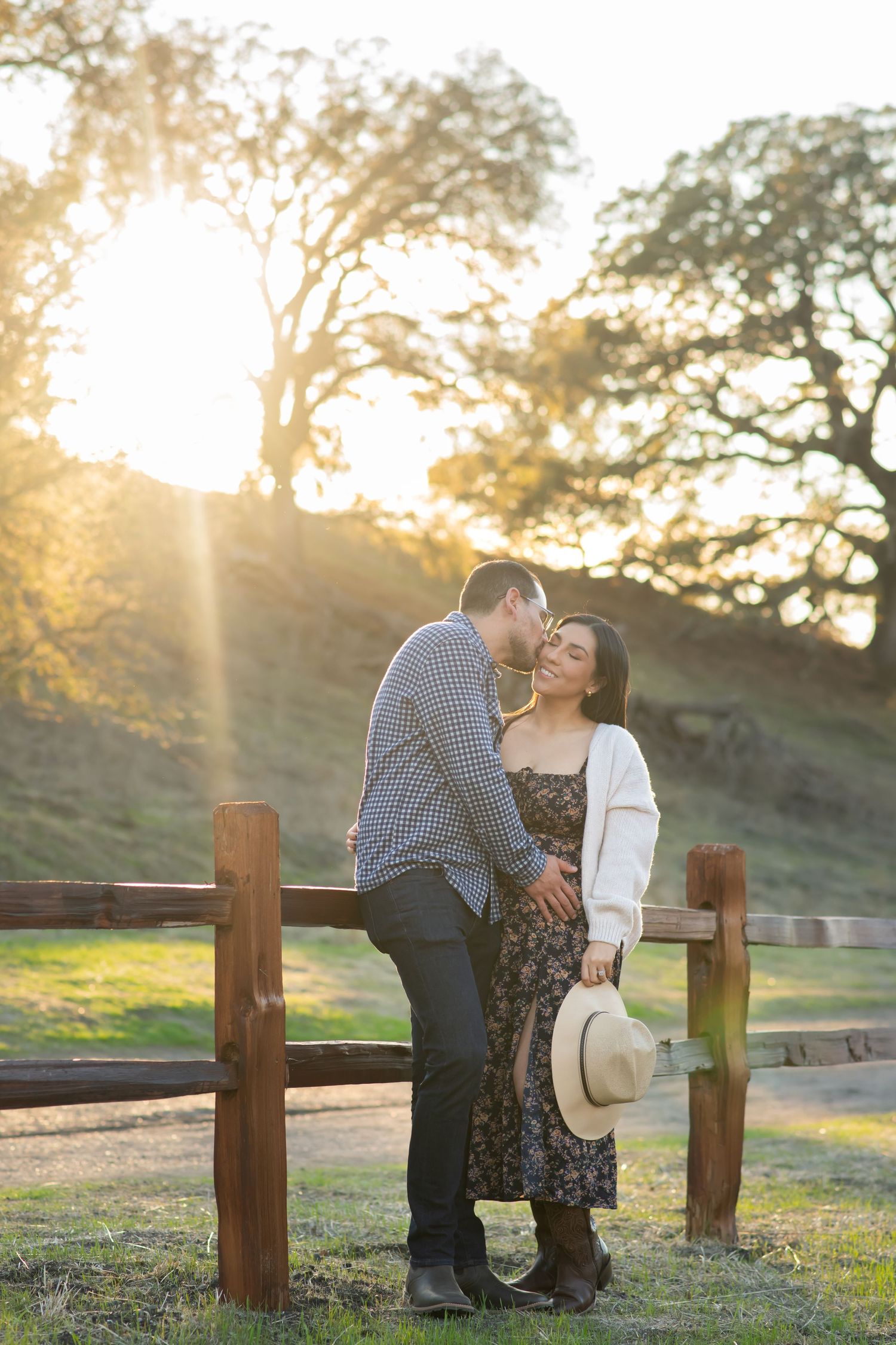 A couple shares a romantic moment at sunset by a wooden fence in a scenic rural setting.