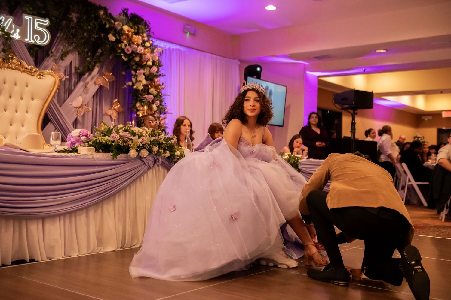 A quinceañera celebration with someone adjusting a flowing white ball gown in front of an elegantly decorated backdrop.
