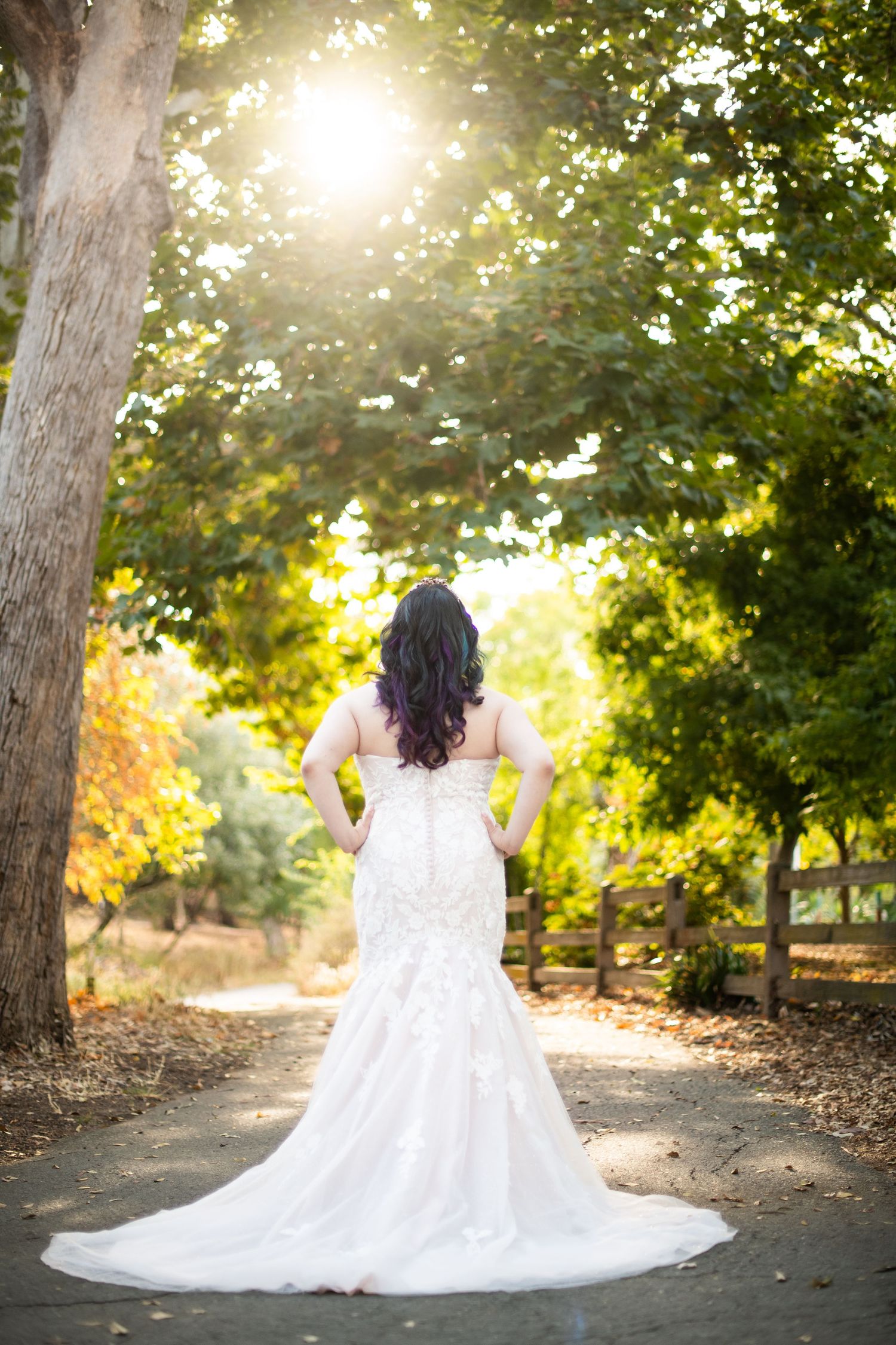 A person in a flowing white wedding dress stands on a sunlit path surrounded by autumn trees.