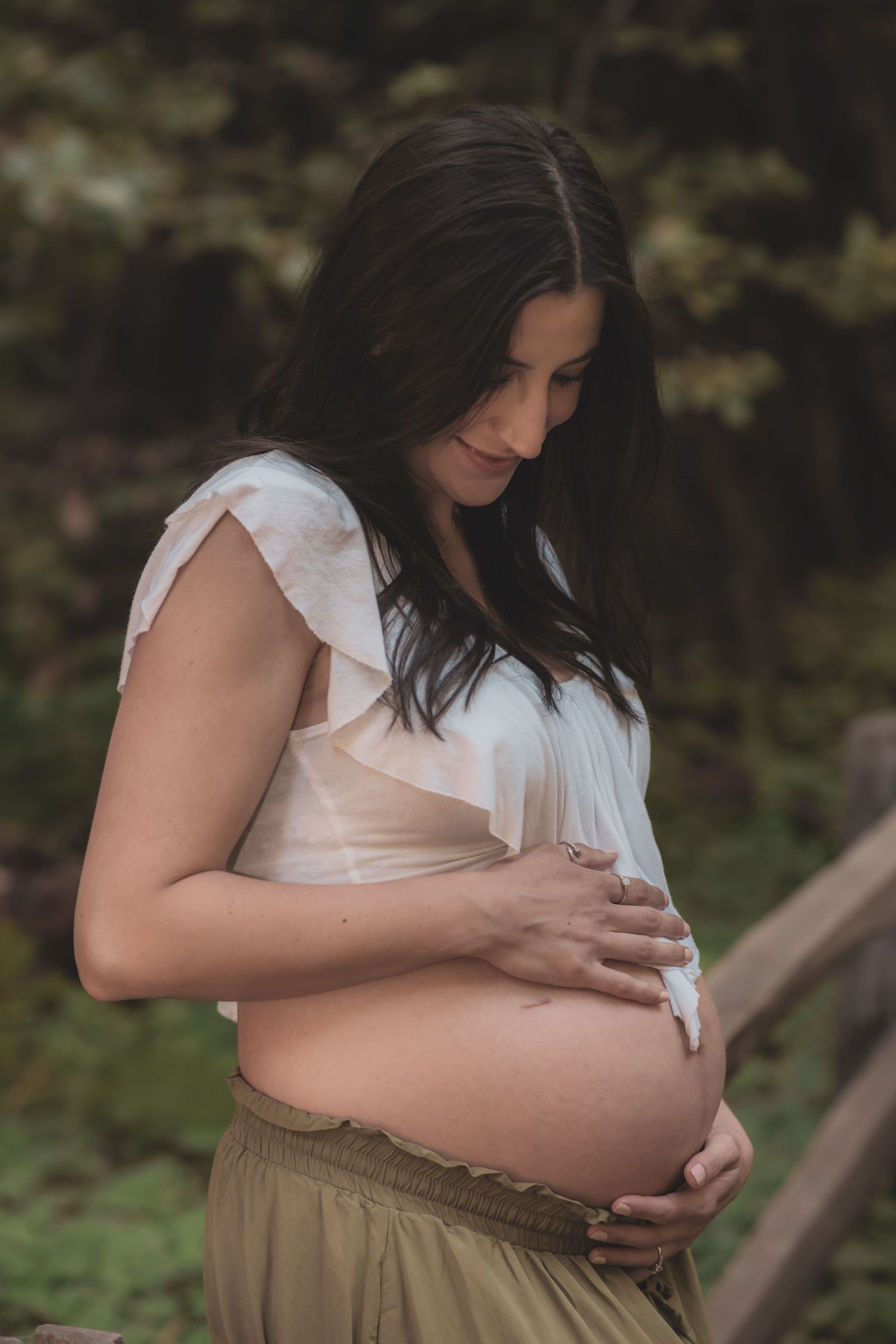 A pregnant woman in a white ruffle top and brown skirt tenderly holds her baby bump in an outdoor nature setting.
