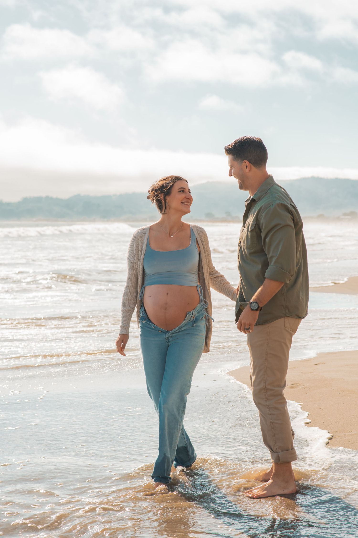 A joyful expectant couple shares a tender moment while walking along the beach shoreline.