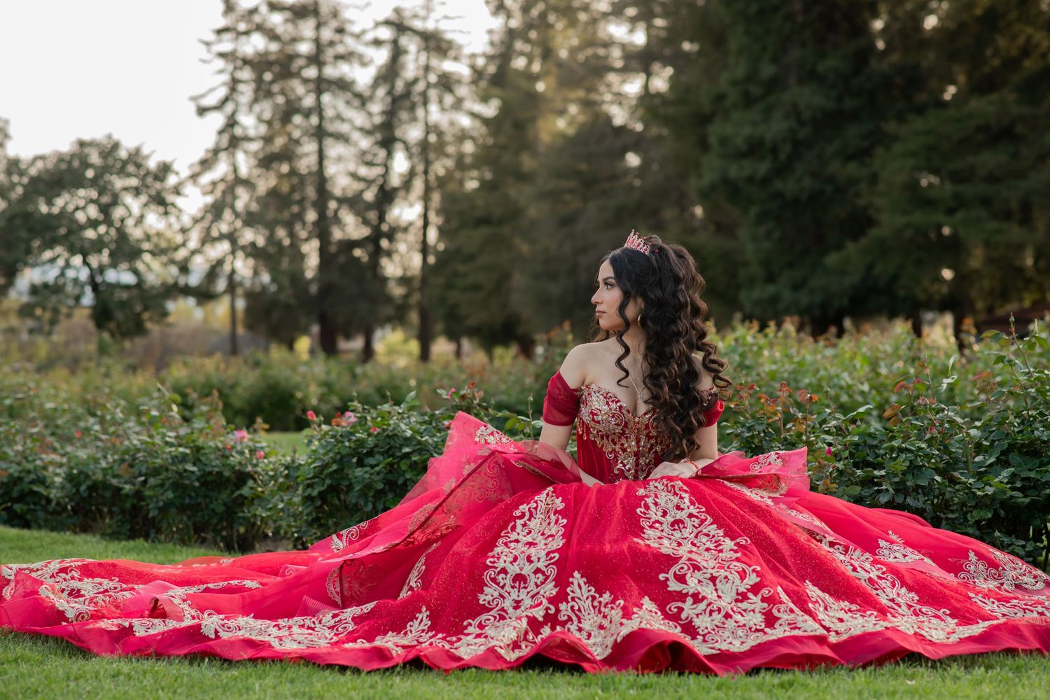 A person in a stunning red and gold embellished gown sits gracefully in a lush garden setting during golden hour.