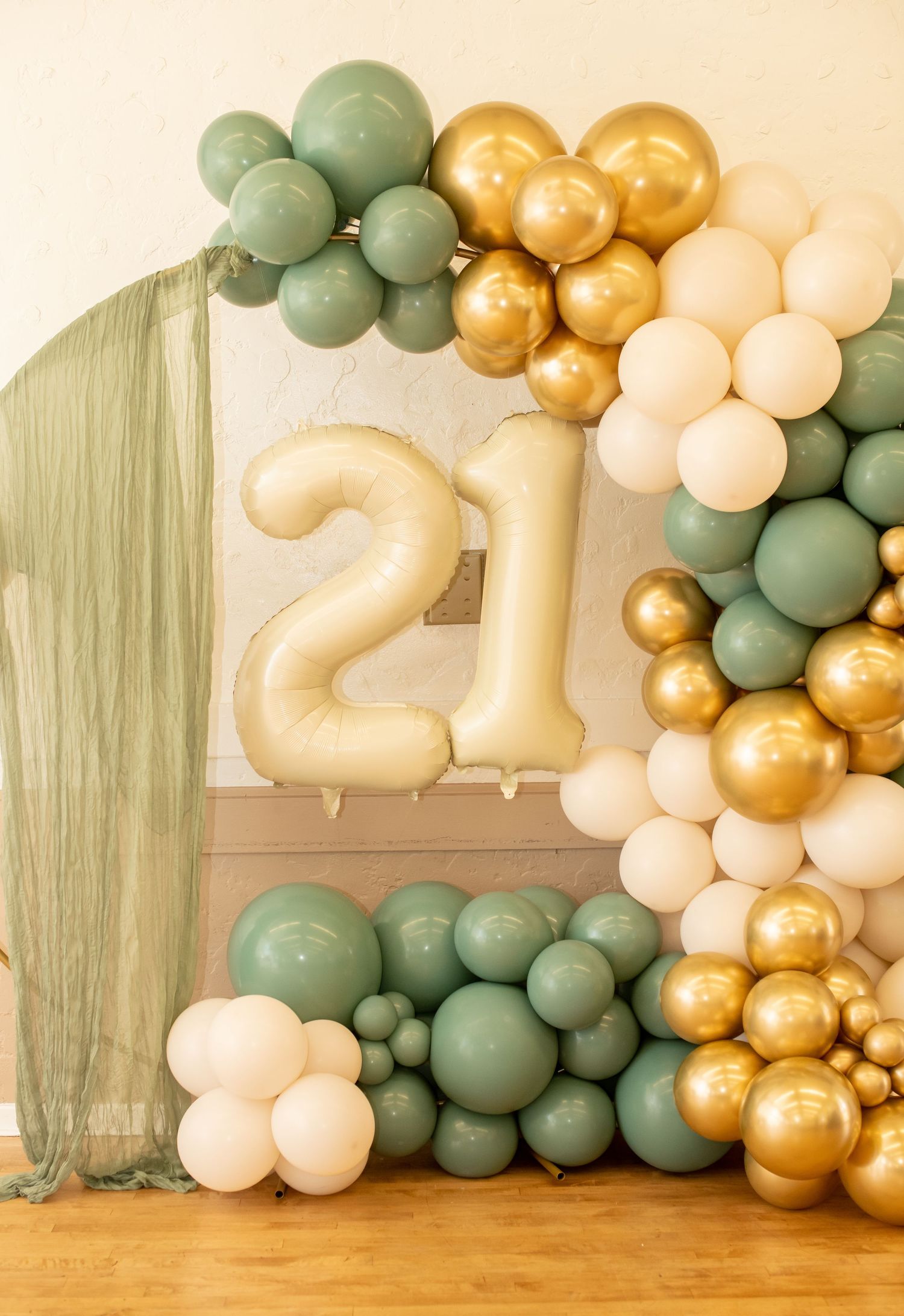 A balloon arch decoration featuring the number 21 in gold with sage green, gold, and cream colored balloons.