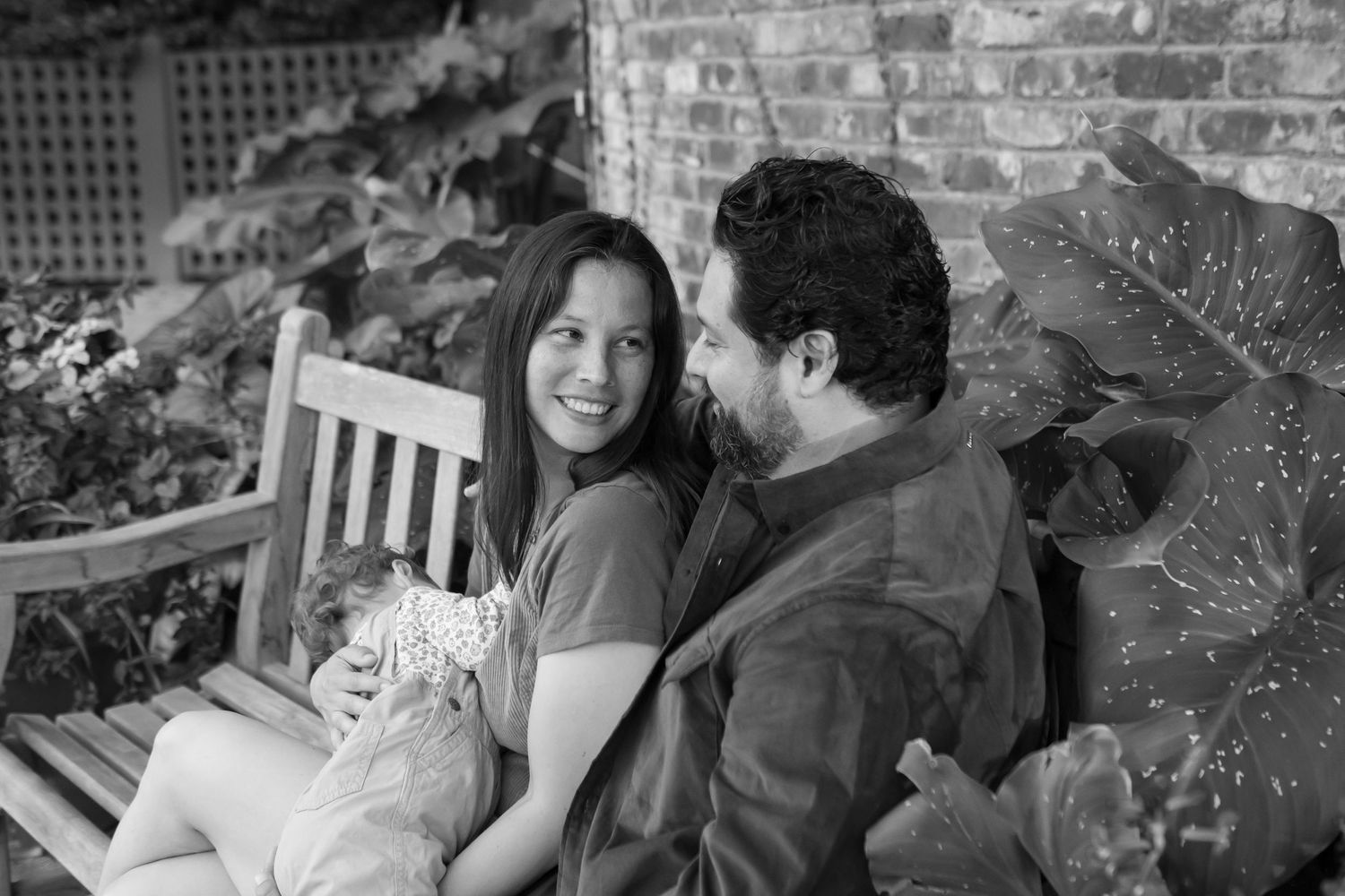 Black and white photo of a couple sitting close together on a wooden bench in front of a brick wall.