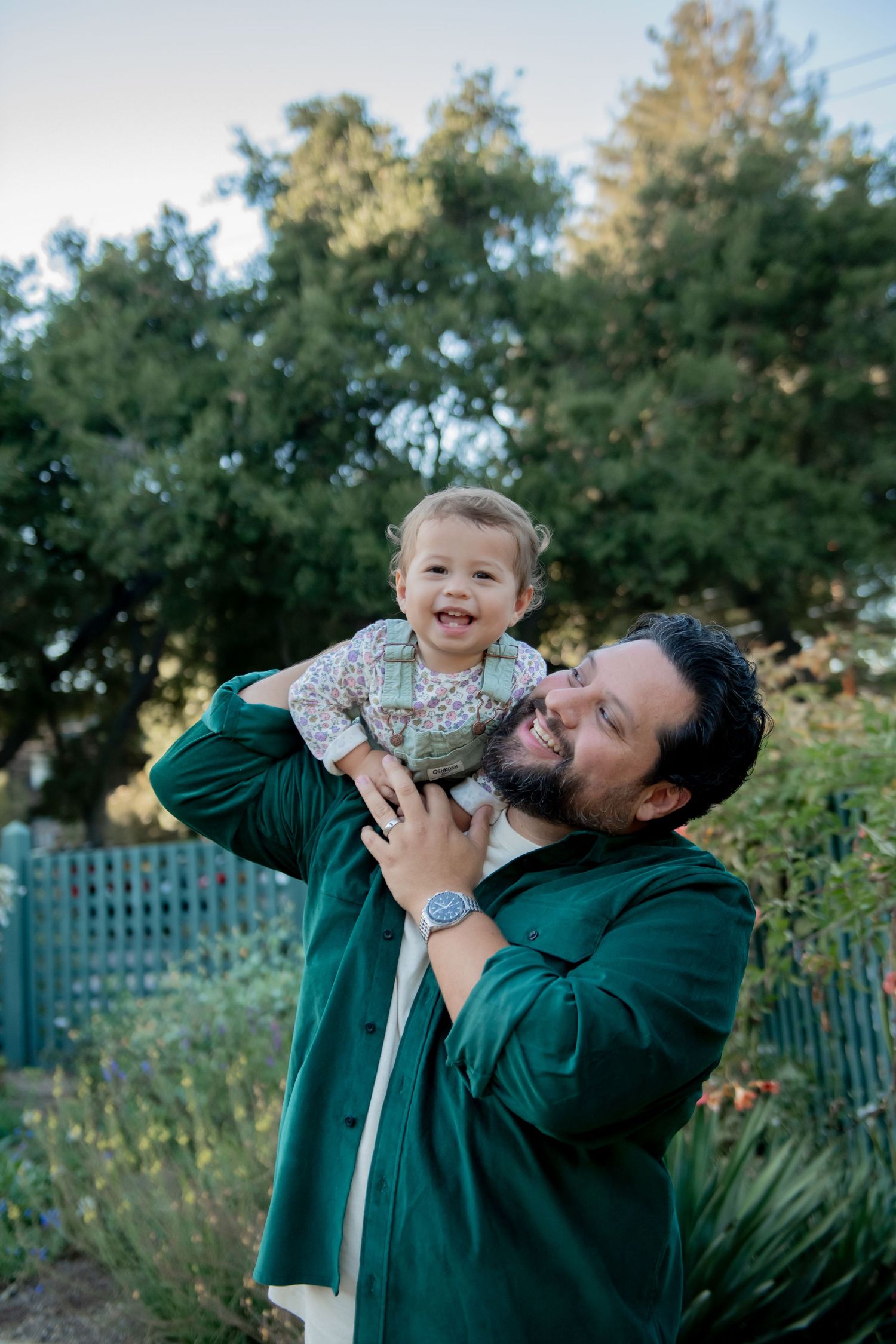 A joyful interaction between two people outdoors on a sunny day in front of lush green trees and garden foliage.