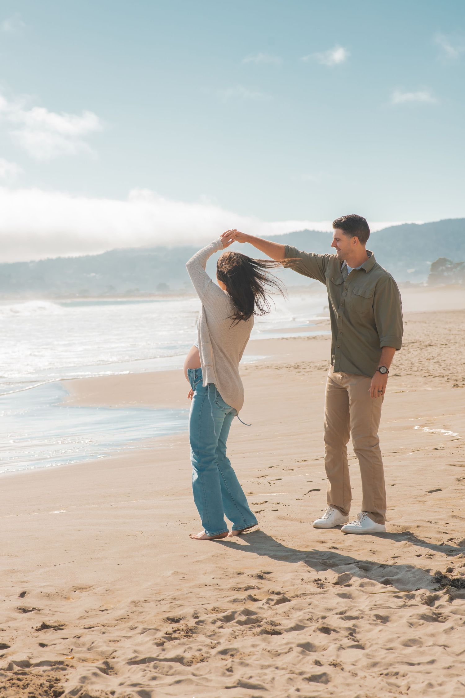 A couple dances together on a scenic beach with mountains and waves in the background.