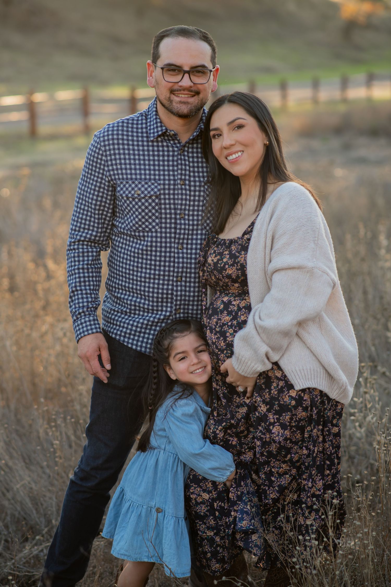 Family of three poses together in a field during golden hour wearing coordinated casual outfits.