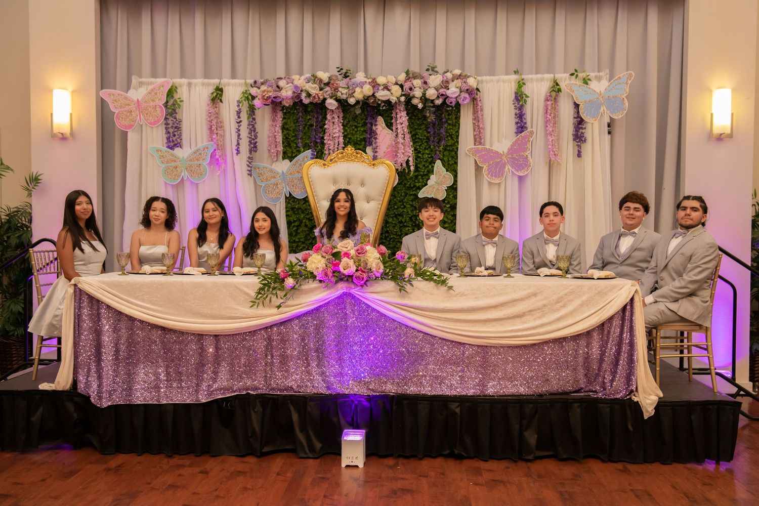 Wedding party seated at an elegant head table decorated with floral arrangements and purple lighting effects.