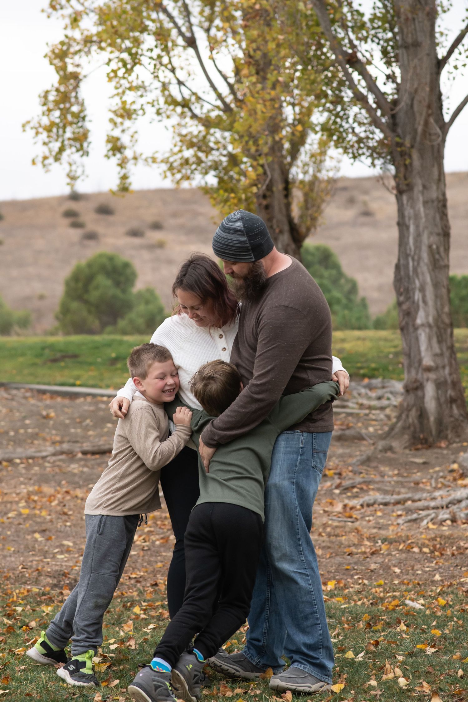 A family shares a warm embrace while standing on autumn leaves beneath tall trees in a rural outdoor setting.
