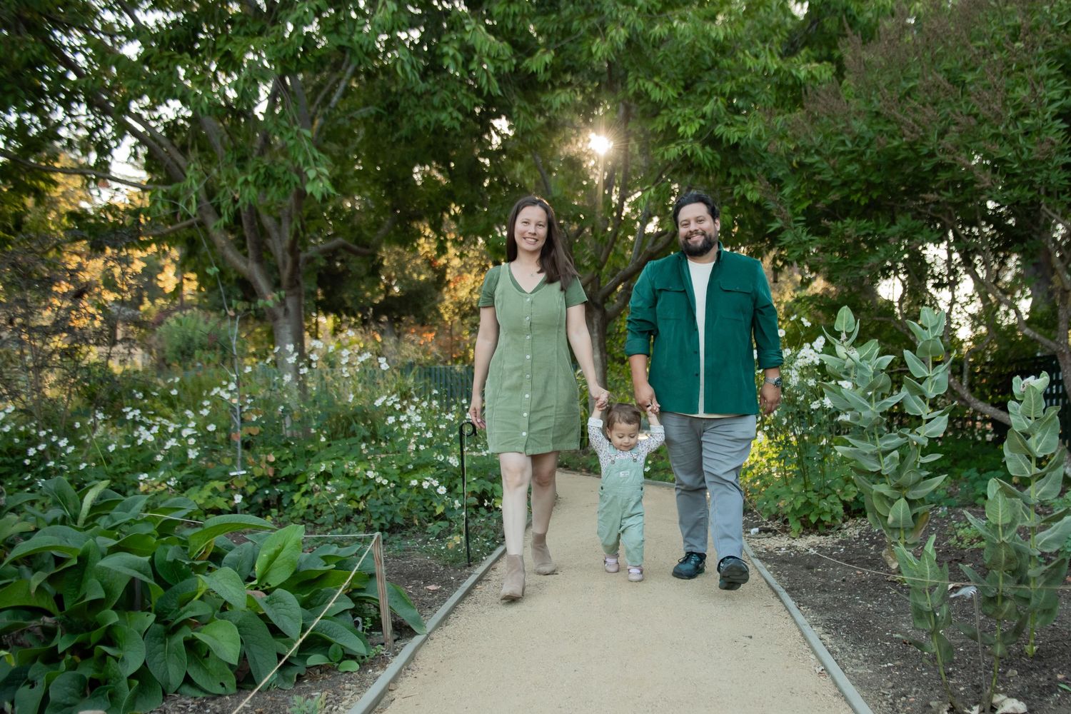 Three people walk together holding hands on a garden path during golden hour with lush greenery surrounding them.