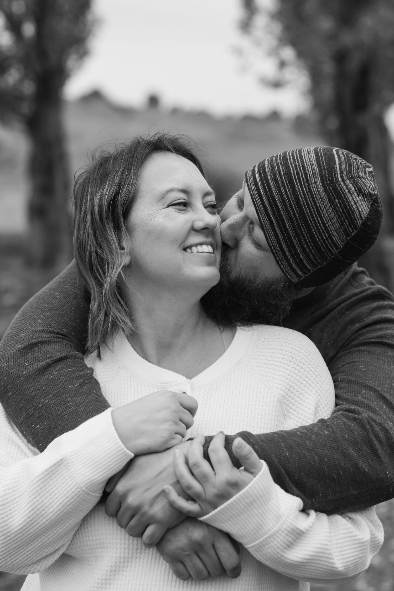 A romantic black and white portrait of a couple sharing a tender embrace outdoors, with one wearing a striped beanie.