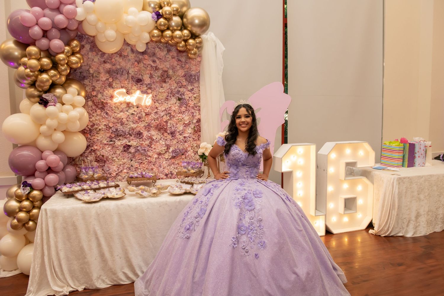 Elegant quinceañera celebration setup with lavender balloons, floral wall backdrop and illuminated numbers in a banquet hall.