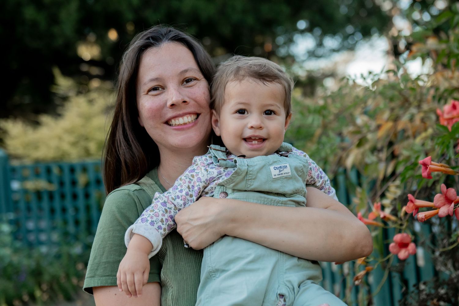 A parent and toddler embrace while smiling outdoors in a garden with flowers and greenery in the background.