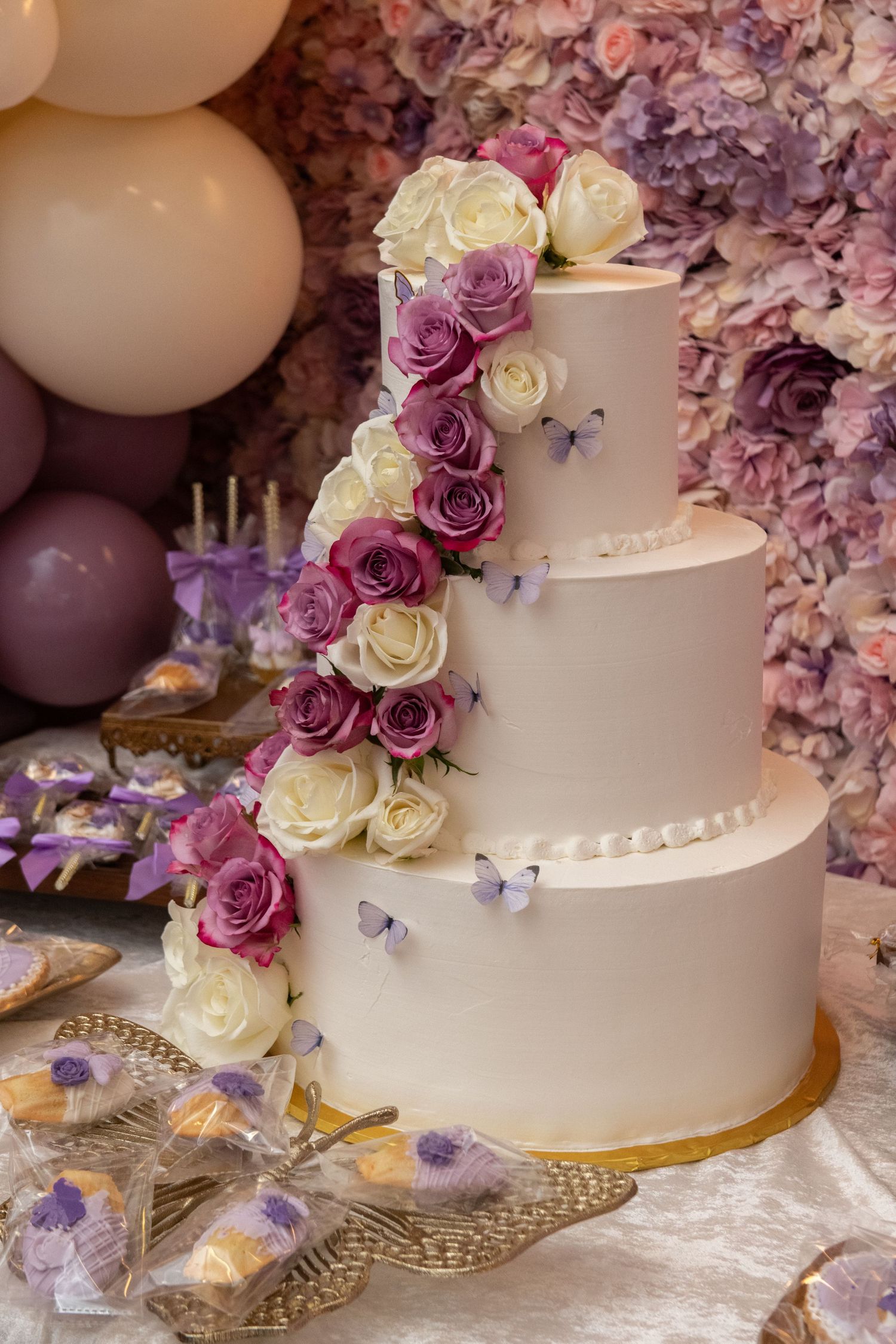 Three-tier white wedding cake decorated with cascading purple and cream roses and butterflies against a floral backdrop.