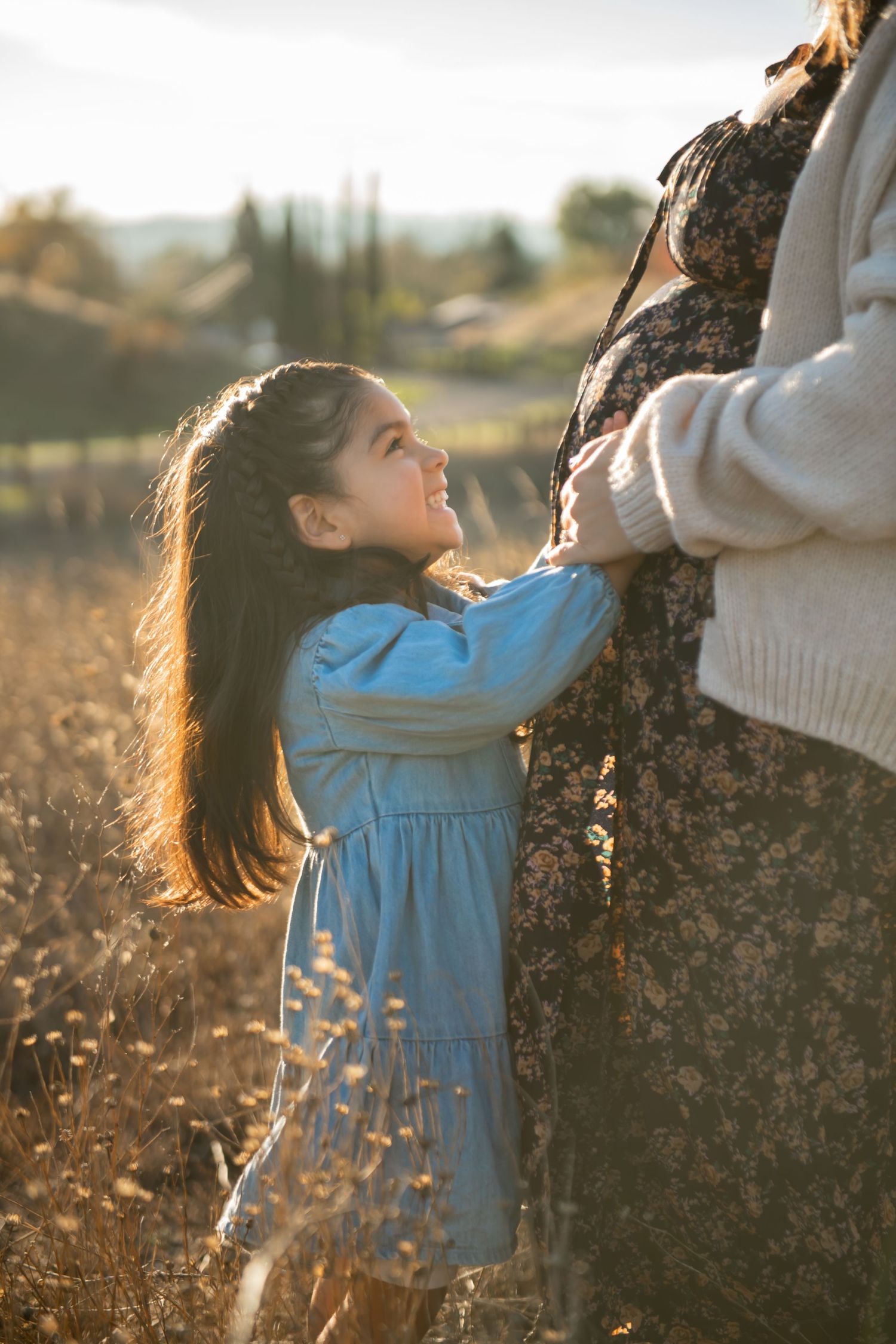 A child in a blue dress reaches up towards a tree trunk against a golden sunset backdrop in a field of tall grass.