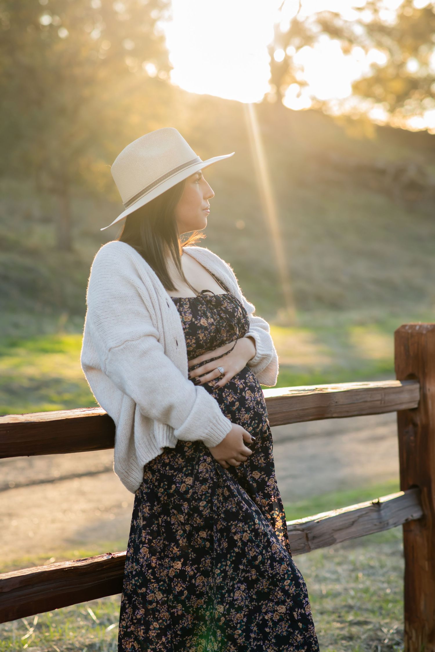 A pregnant woman in a floral dress and sunhat stands by a wooden fence during golden sunset hour.