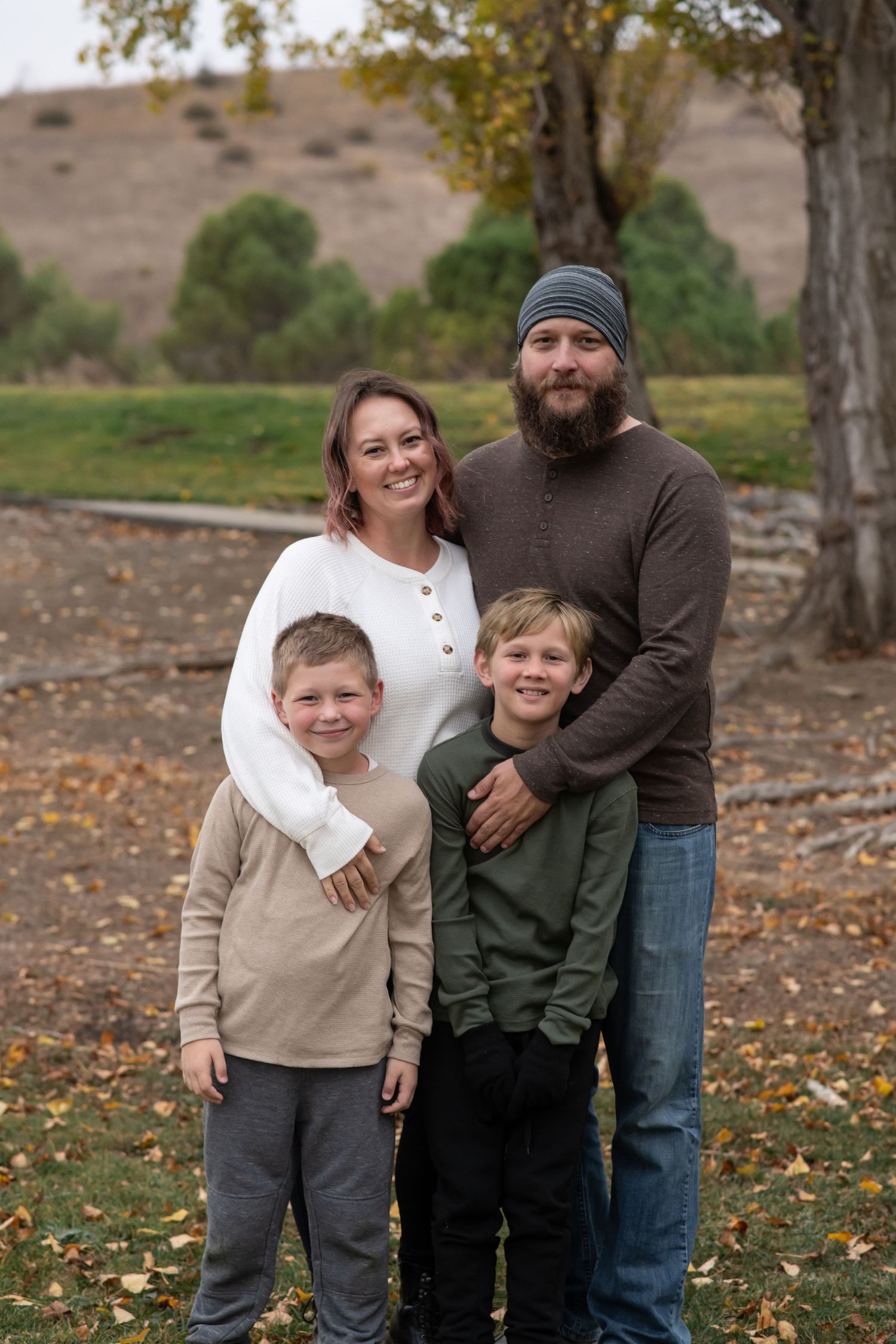 Family of four wearing casual fall clothing poses together on an autumn day with trees and hills in the background.