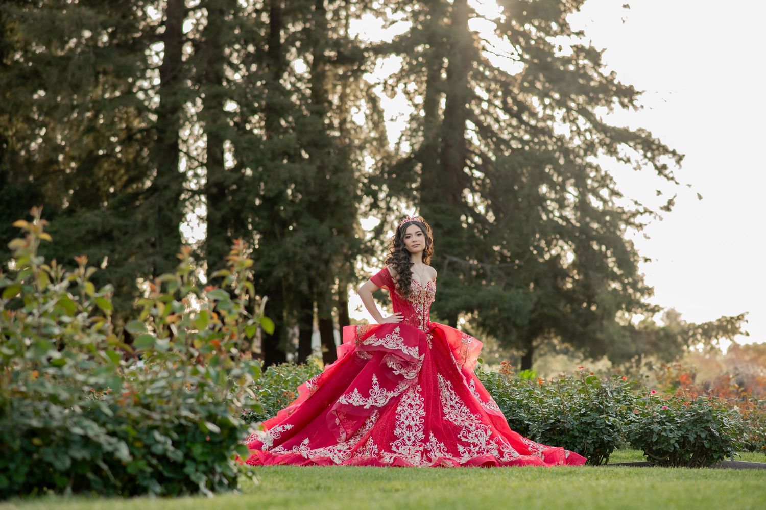 A stunning red ballgown with intricate embroidery sparkles in a sunlit garden surrounded by tall evergreen trees.