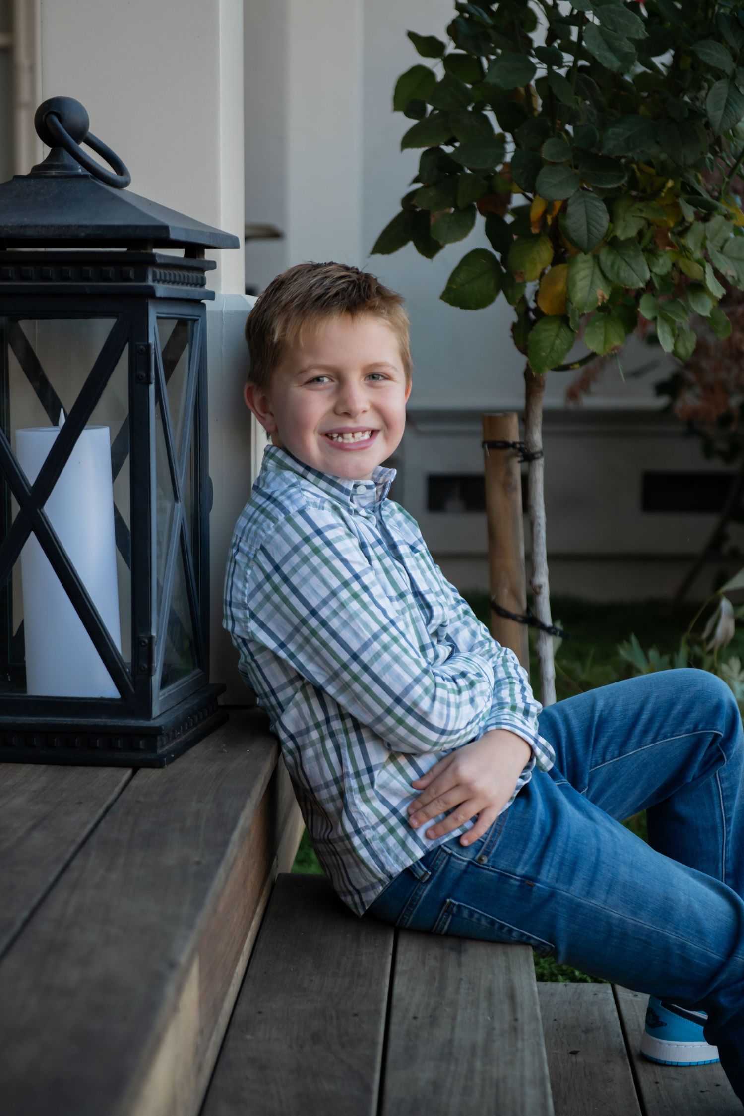 Young person in plaid shirt and jeans sits casually on wooden deck with decorative lantern and greenery in background.