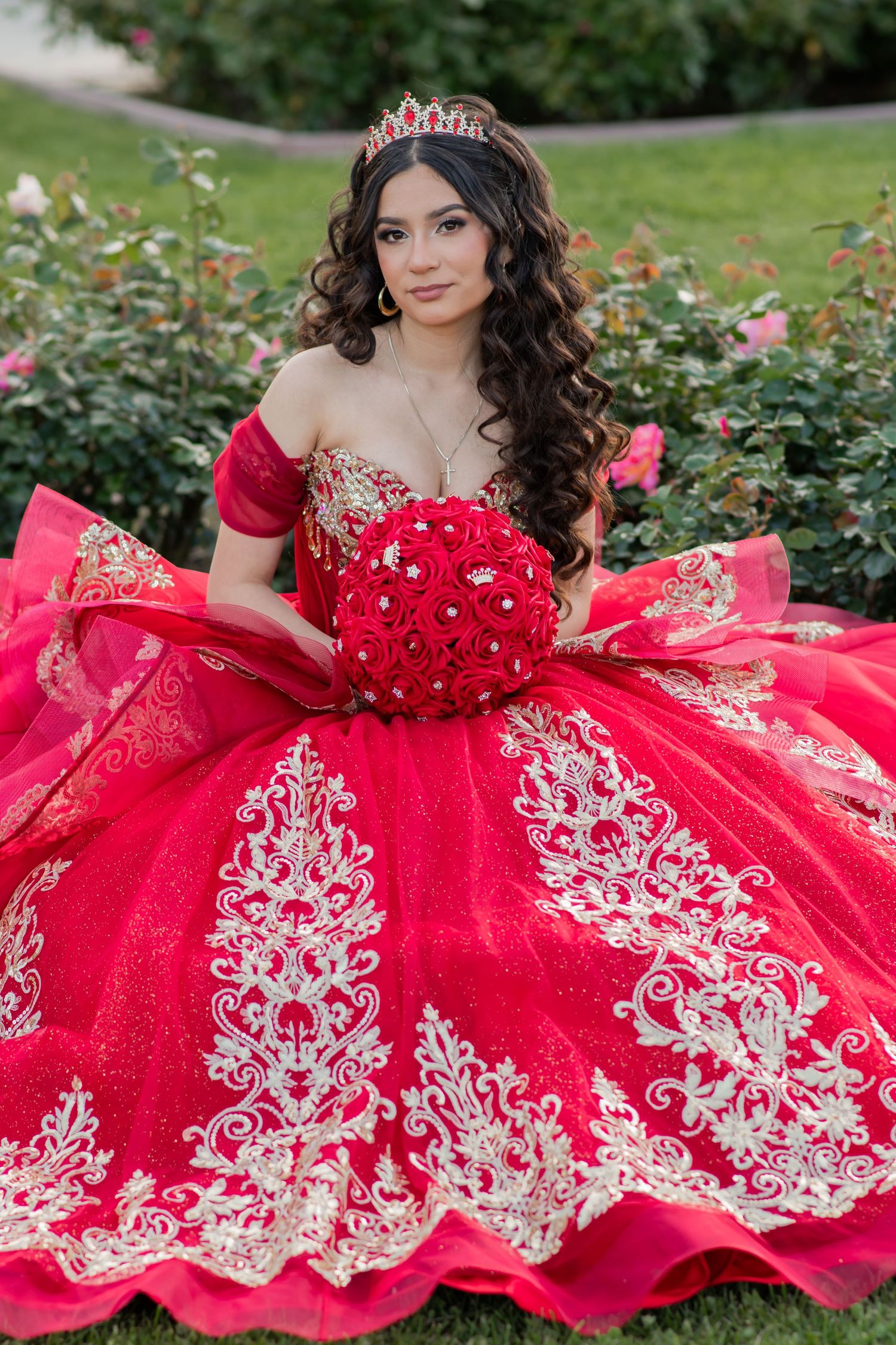 Beautiful red quinceañera dress with ornate white lace embroidery and off-shoulder design photographed in garden setting.