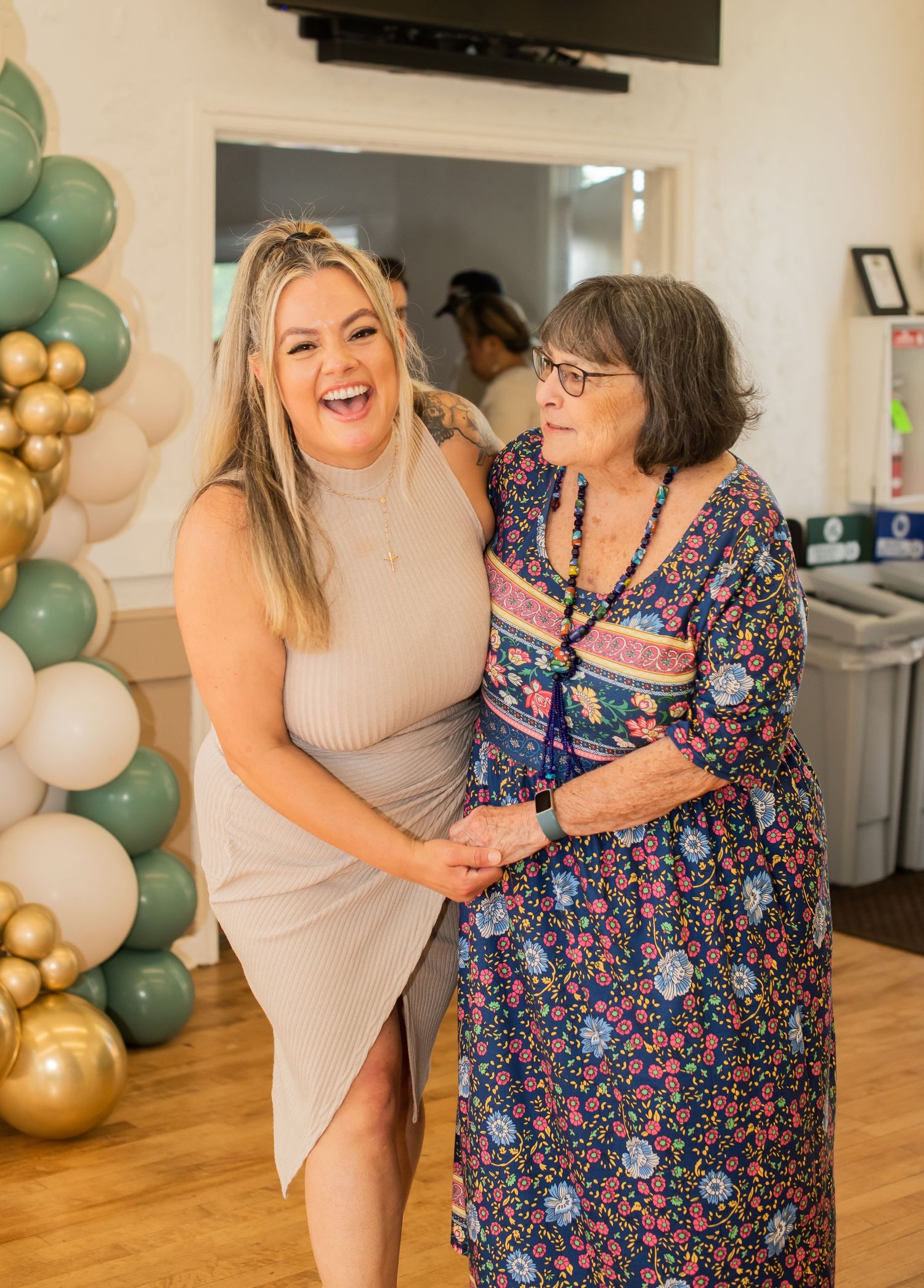 Two people share a joyful moment in front of a festive balloon arch decorated in green, gold and white colors.