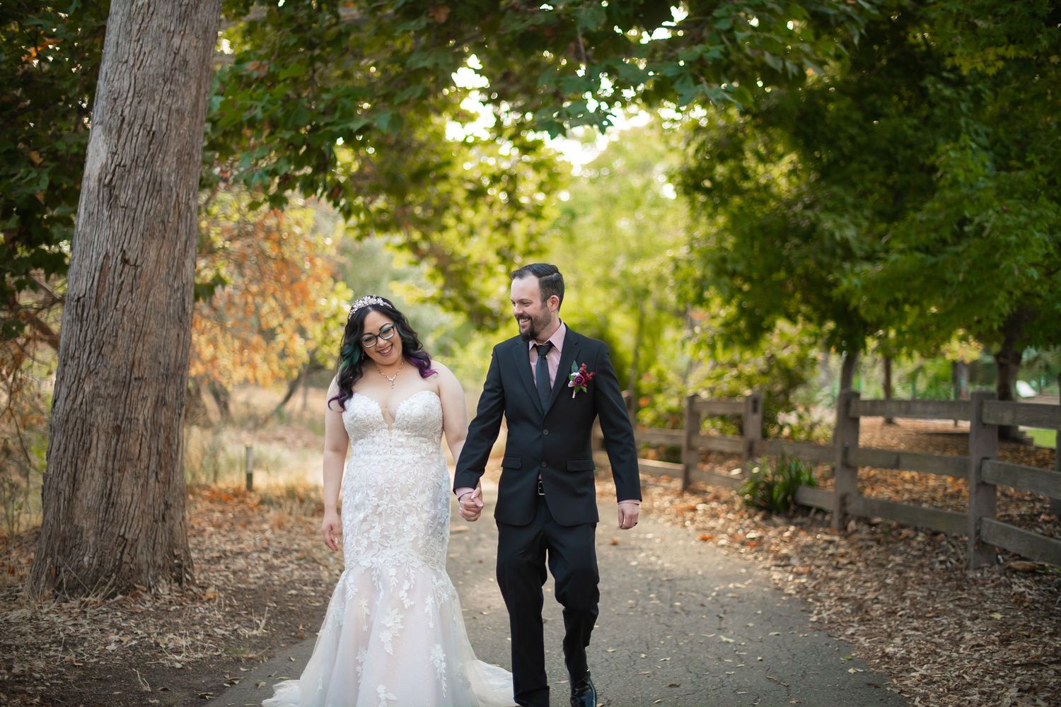 Newlyweds walk hand in hand down a tree-lined path in an autumn park wearing elegant wedding attire.
