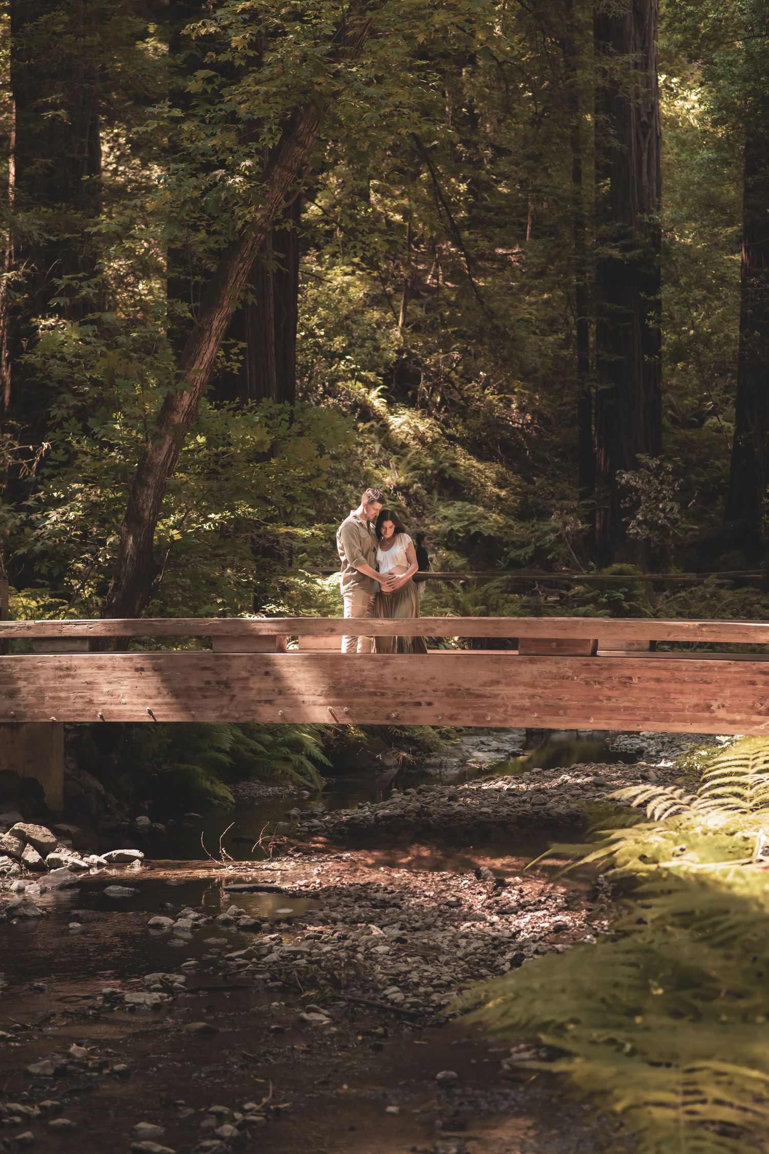 A romantic couple embraces on a wooden footbridge spanning a peaceful forest stream in dappled sunlight.