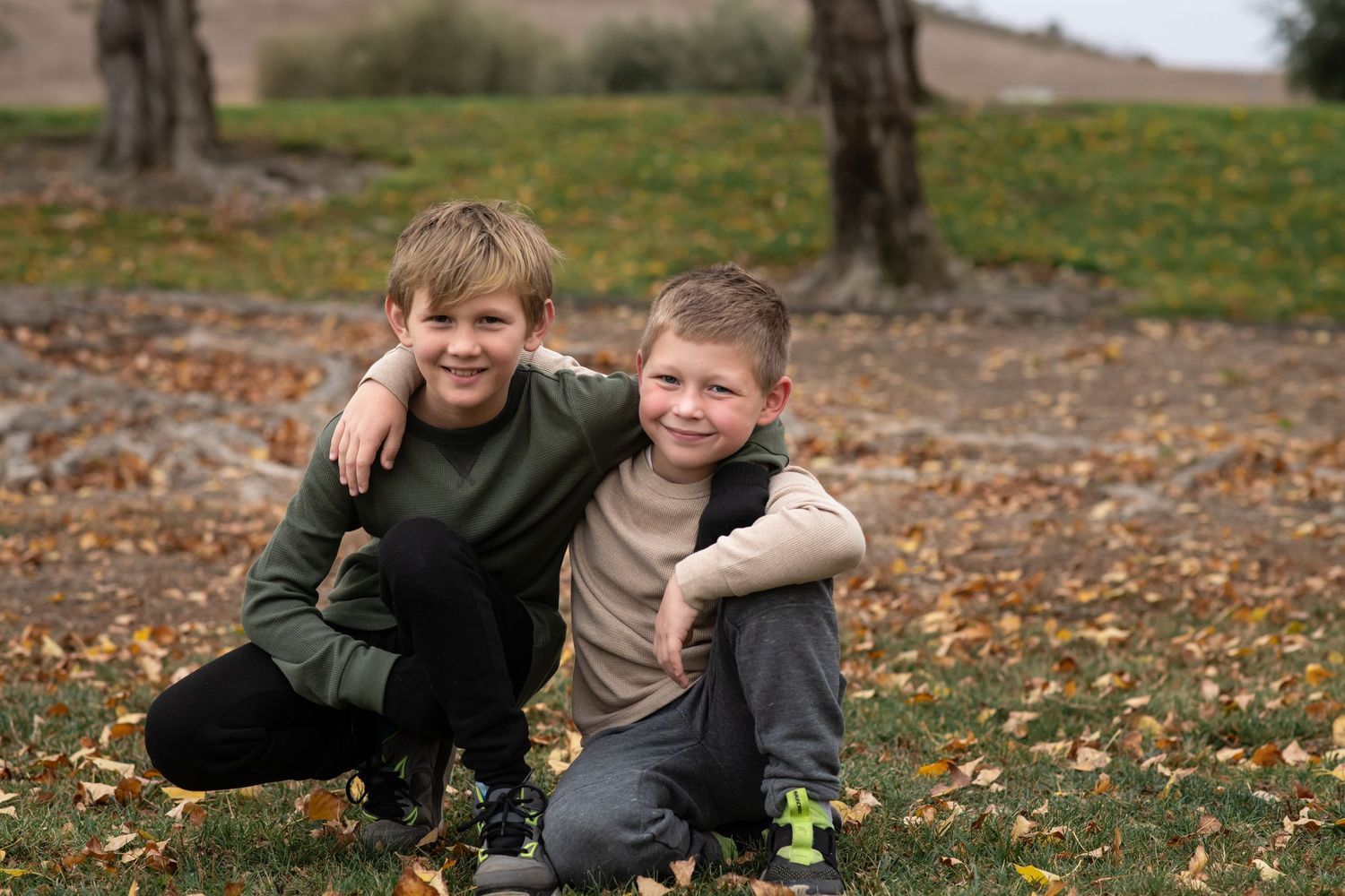 Two children sit close together on autumn ground covered in fallen leaves and dandelions.