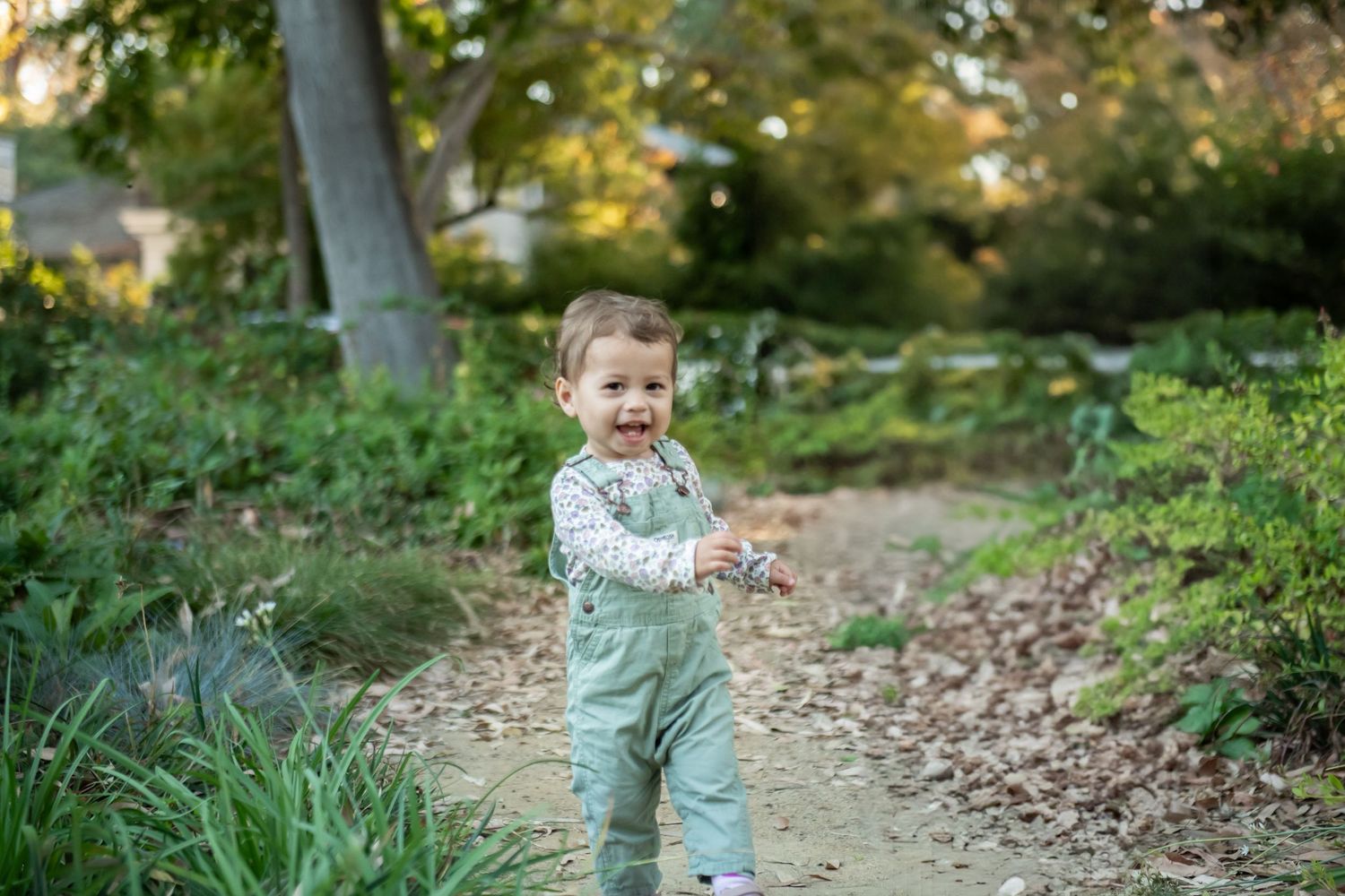 Small child wearing mint green overalls stands on a wooded path surrounded by autumn leaves and greenery.