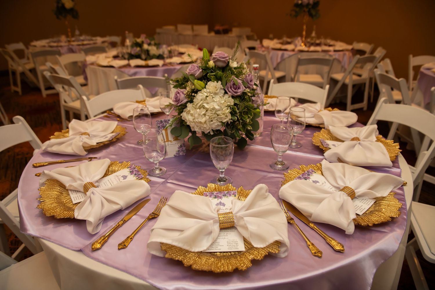 Elegant wedding reception table setting with white napkins, gold chargers, and purple linens centered by a hydrangea floral arrangement.