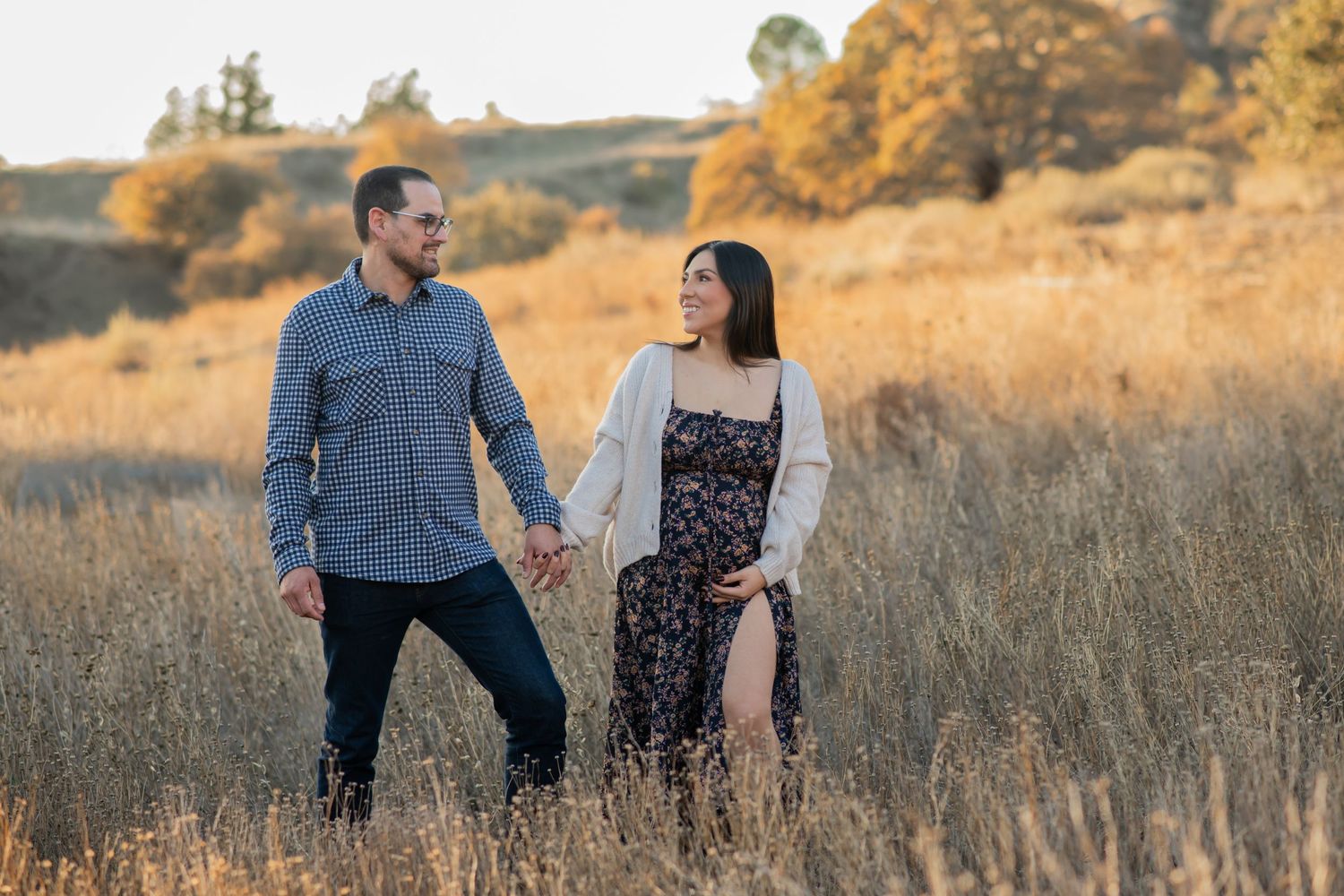 A couple holds hands while walking through a golden wheat field during sunset in a romantic outdoor setting.